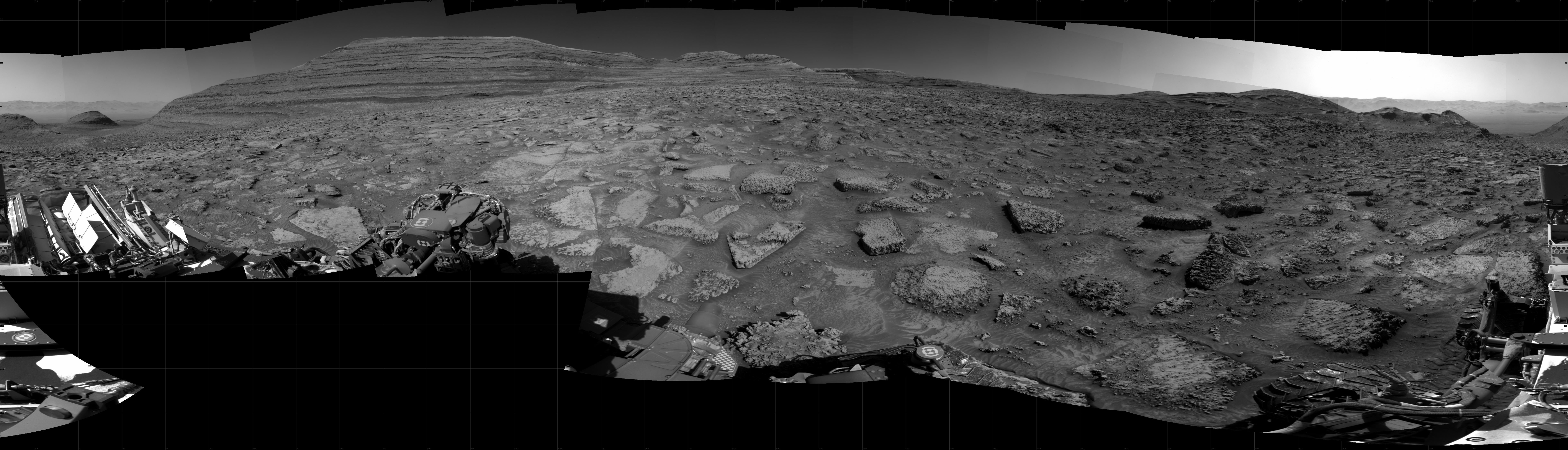 Panoramic black-and-white image of the rocky Martian landscape taken by the Curiosity rover, showing rugged terrain, layered hills in the distance, and parts of the rover's structure in the foreground.