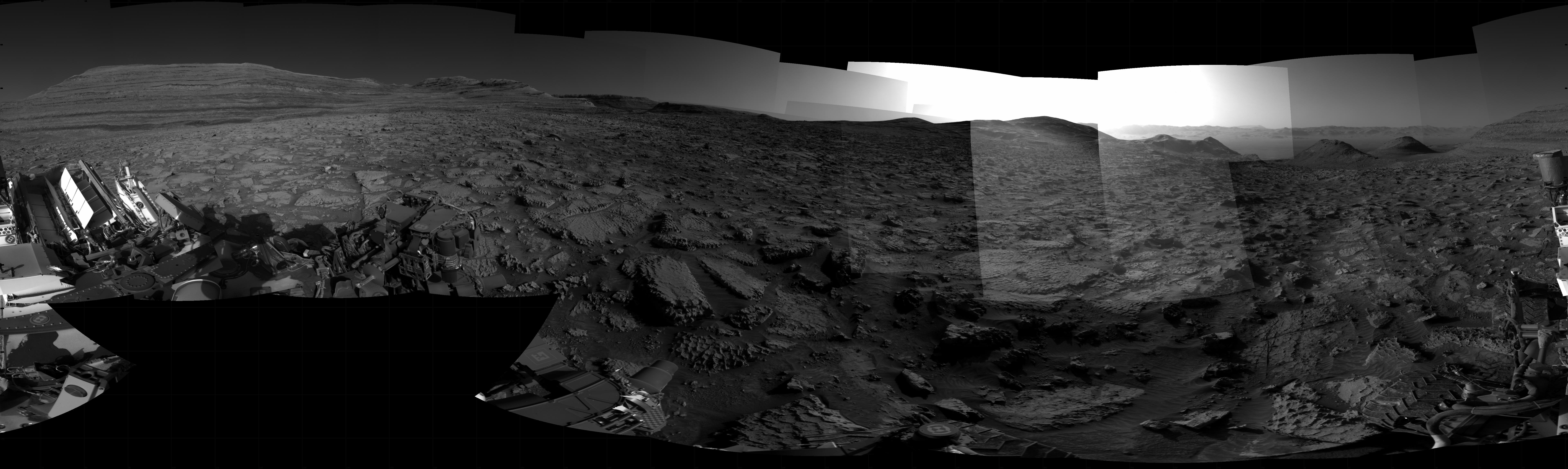 Panoramic black-and-white image of the rocky Martian landscape taken by the Curiosity rover, showing rugged terrain, layered hills in the distance, and parts of the rover's structure in the foreground.