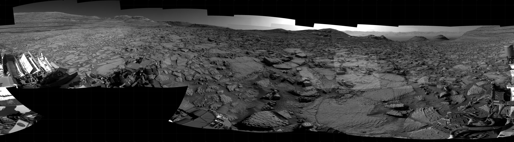 Panoramic black-and-white image of the rocky Martian landscape taken by the Curiosity rover, showing rugged terrain, layered hills in the distance, and parts of the rover's structure in the foreground.