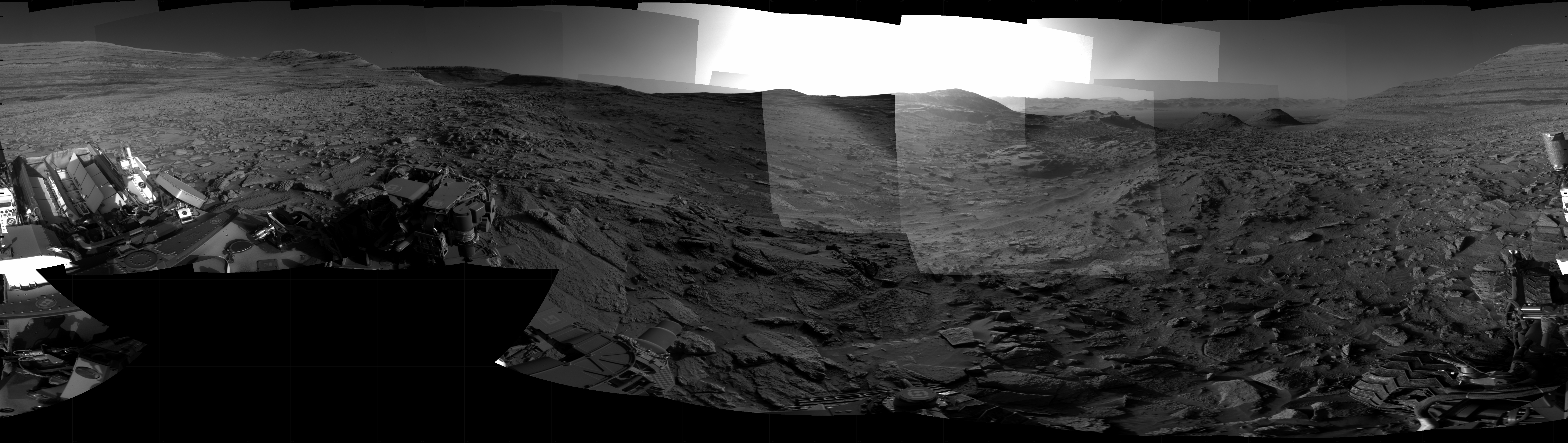 Panoramic black-and-white image of the rocky Martian landscape taken by the Curiosity rover, showing rugged terrain, layered hills in the distance, and parts of the rover's structure in the foreground.
