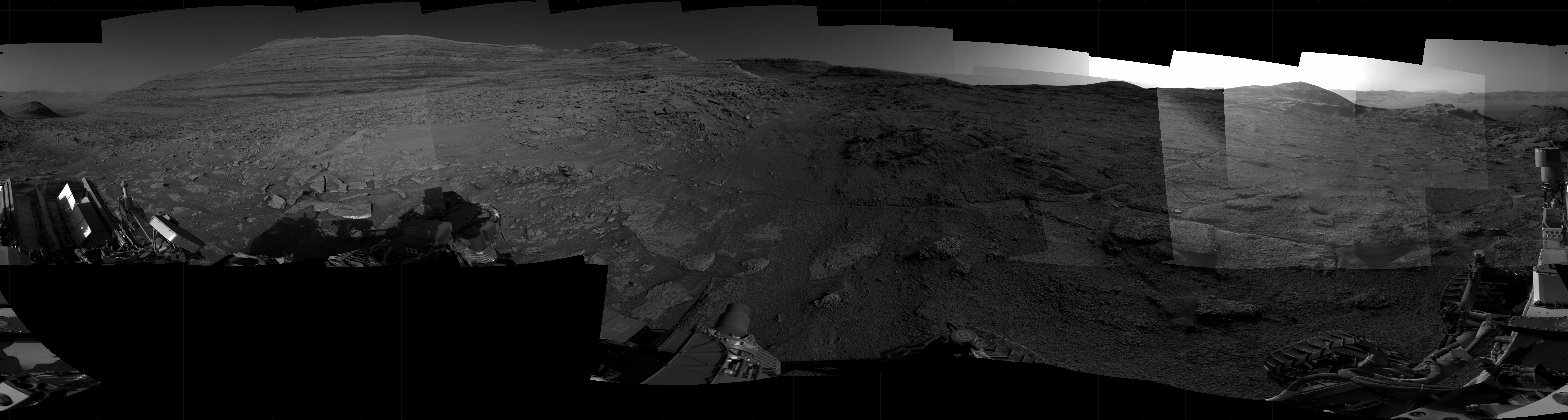 At the far right of the frame, a very bright light source illuminates the sky above several small hills. Portions of the Curiosity rover are visible along the bottom of the image, including one wheel visible in the bottom center of the frame and two others in the lower right corner.