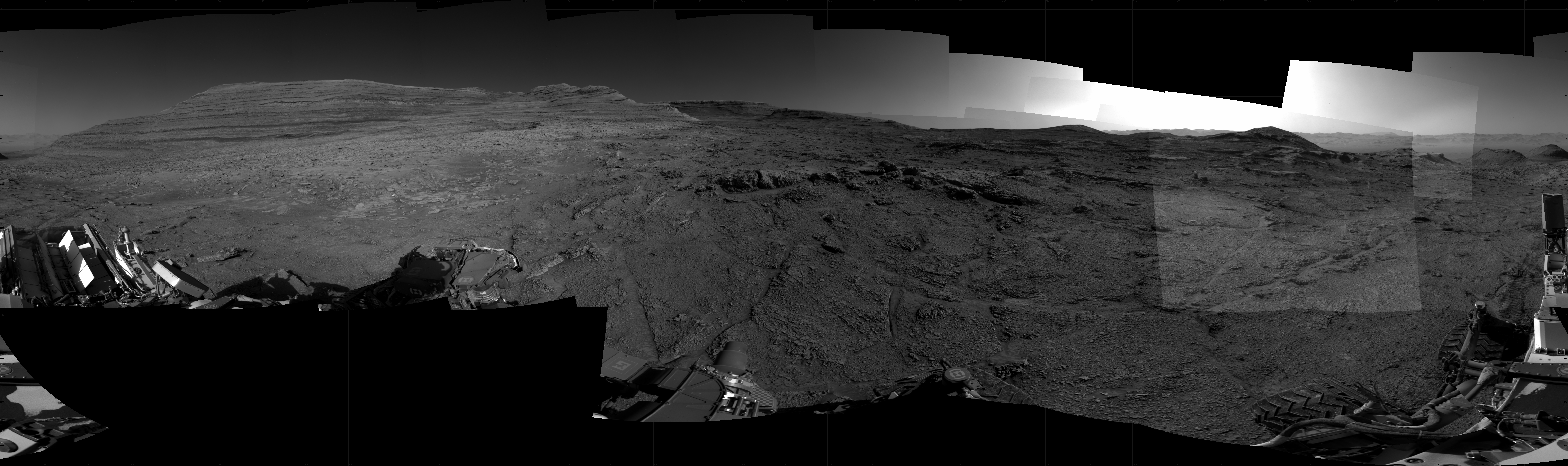 A grayscale panorama of the Martian surface, possibly at night or in twilight, shows a wide field of flat, dark gray terrain dotted with flat, angular, medium-sized rocks stretching into the distance where features rise from the ground. At the far right of the frame, a very bright light source illuminates the sky above several small hills. Portions of the Curiosity rover are visible along the bottom of the image, including one wheel visible in the bottom center of the frame and two others in the lower right corner.