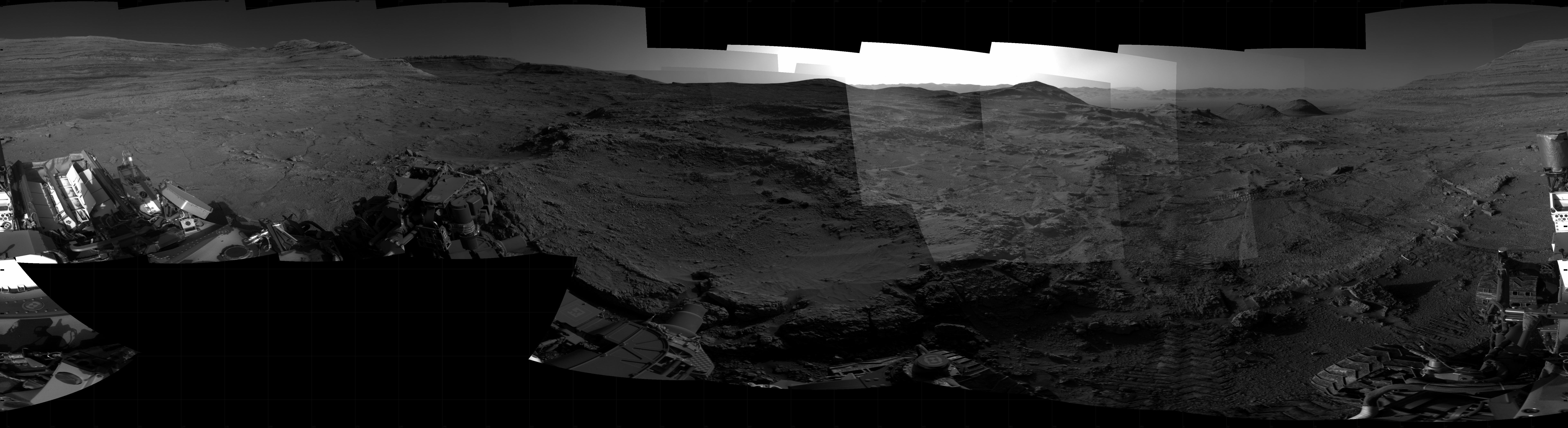 Panoramic black-and-white image of the rocky Martian landscape taken by the Curiosity rover, showing rugged terrain, layered hills in the distance, and parts of the rover's structure in the foreground.