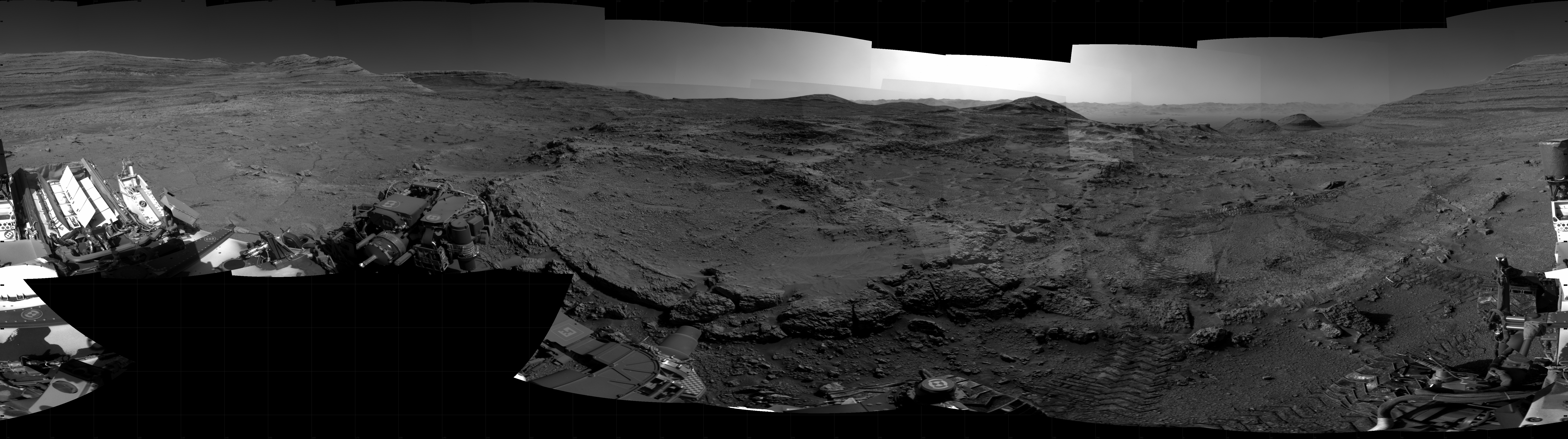Panoramic black-and-white image of the rocky Martian landscape taken by the Curiosity rover, showing rugged terrain, layered hills in the distance, and parts of the rover's structure in the foreground.