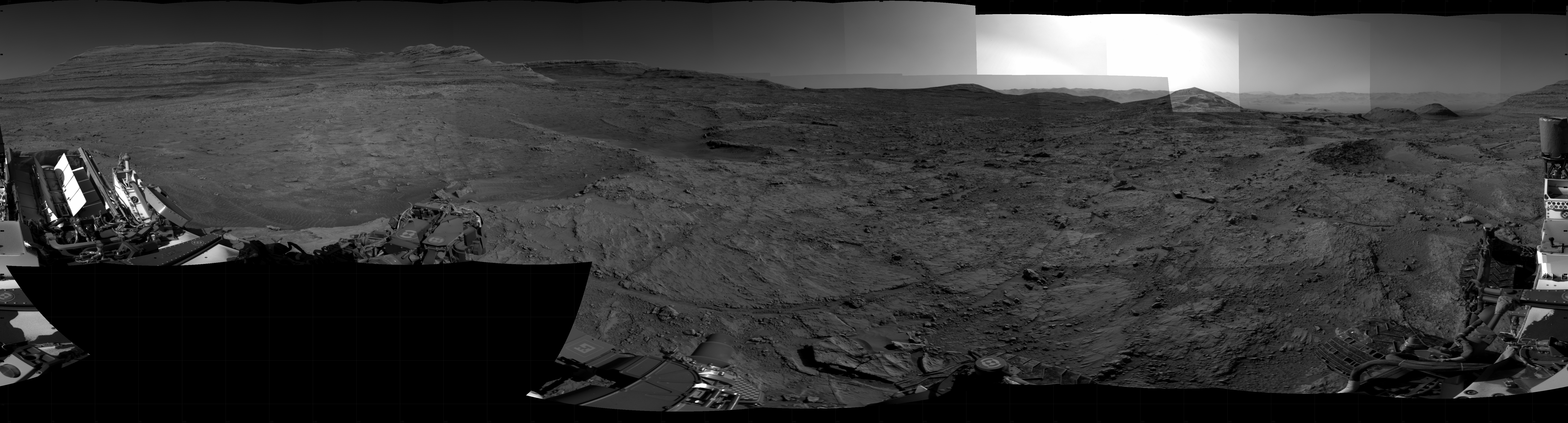 Panoramic black-and-white image of the rocky Martian landscape taken by the Curiosity rover, showing rugged terrain, layered hills in the distance, and parts of the rover's structure in the foreground.