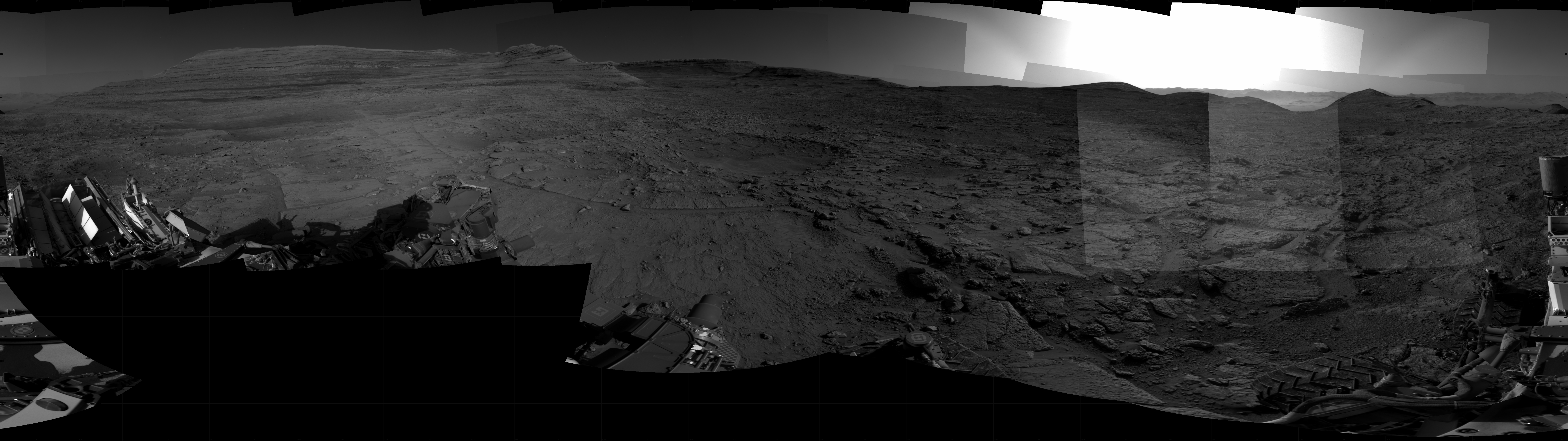 Panoramic black-and-white image of the rocky Martian landscape taken by the Curiosity rover, showing rugged terrain, layered hills in the distance, and parts of the rover's structure in the foreground.