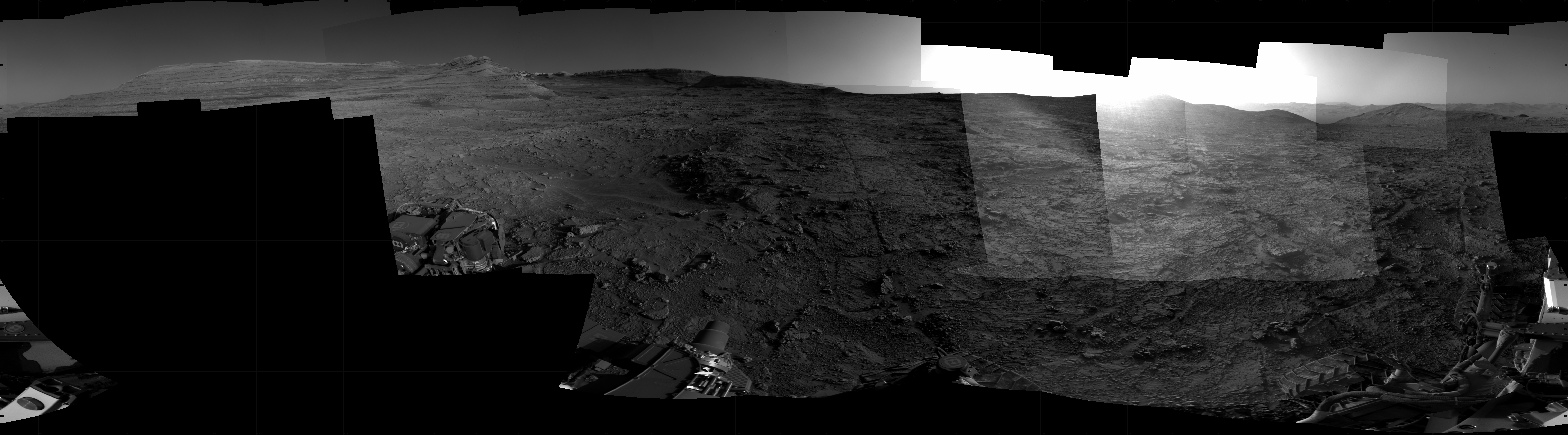 Panoramic black-and-white image of the rocky Martian landscape taken by the Curiosity rover, showing rugged terrain, layered hills in the distance, and parts of the rover's structure in the foreground.
