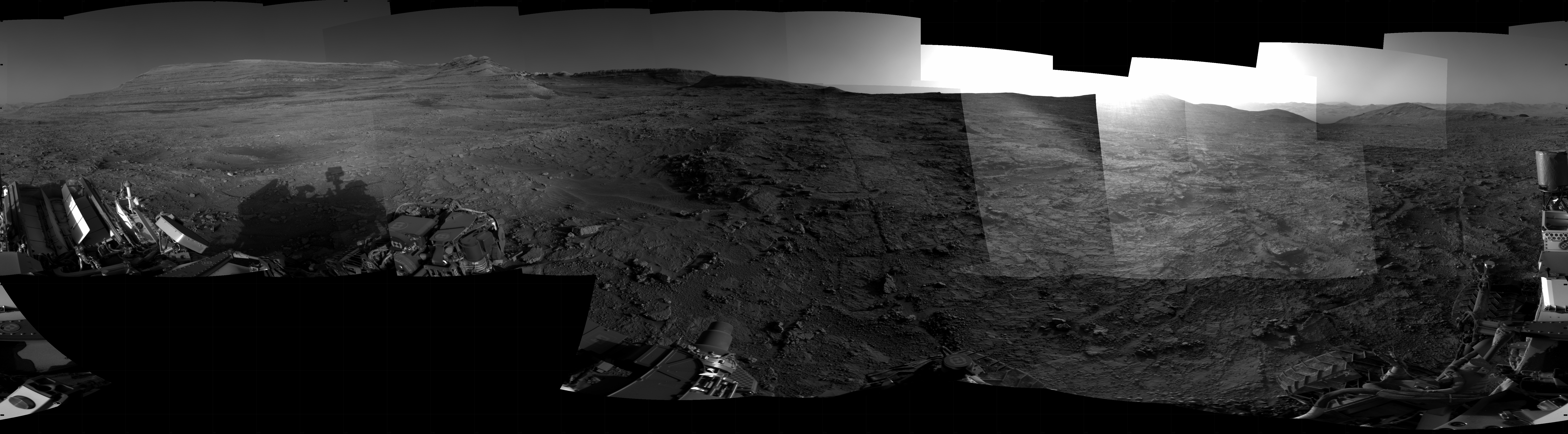 Panoramic black-and-white image of the rocky Martian landscape taken by the Curiosity rover, showing rugged terrain, layered hills in the distance, and parts of the rover's structure in the foreground.