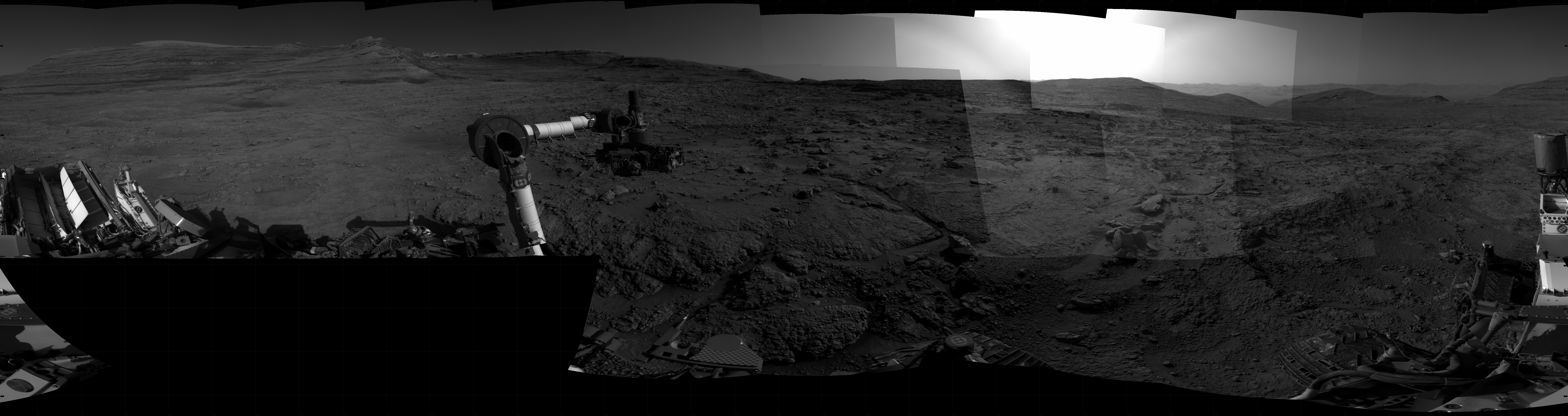 Panoramic black-and-white image of the rocky Martian landscape taken by the Curiosity rover, showing rugged terrain, layered hills in the distance, and parts of the rover's structure in the foreground.