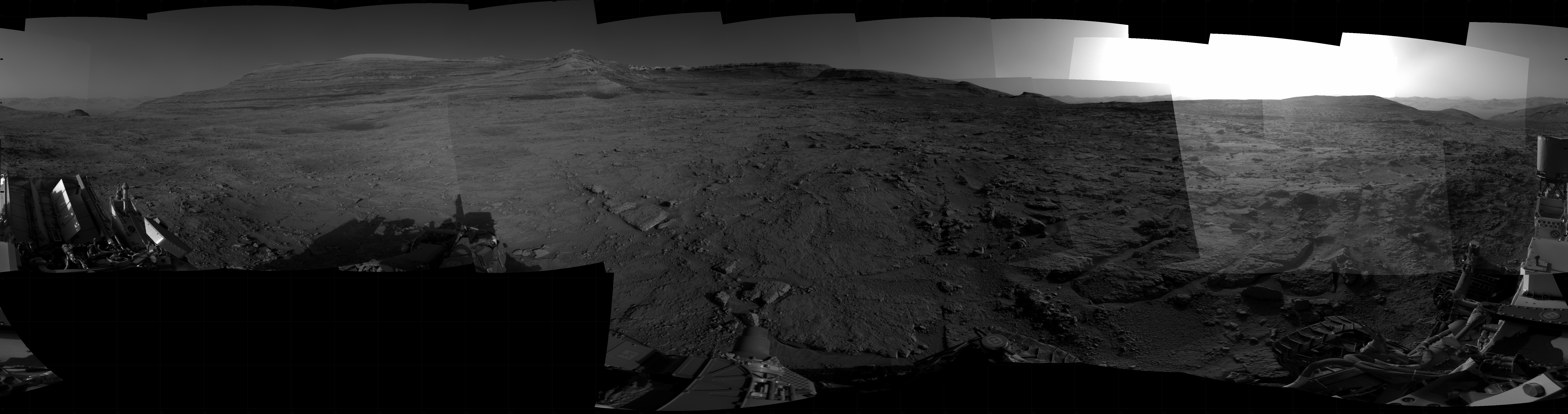 Panoramic black-and-white image of the rocky Martian landscape taken by the Curiosity rover, showing rugged terrain, layered hills in the distance, and parts of the rover's structure in the foreground.