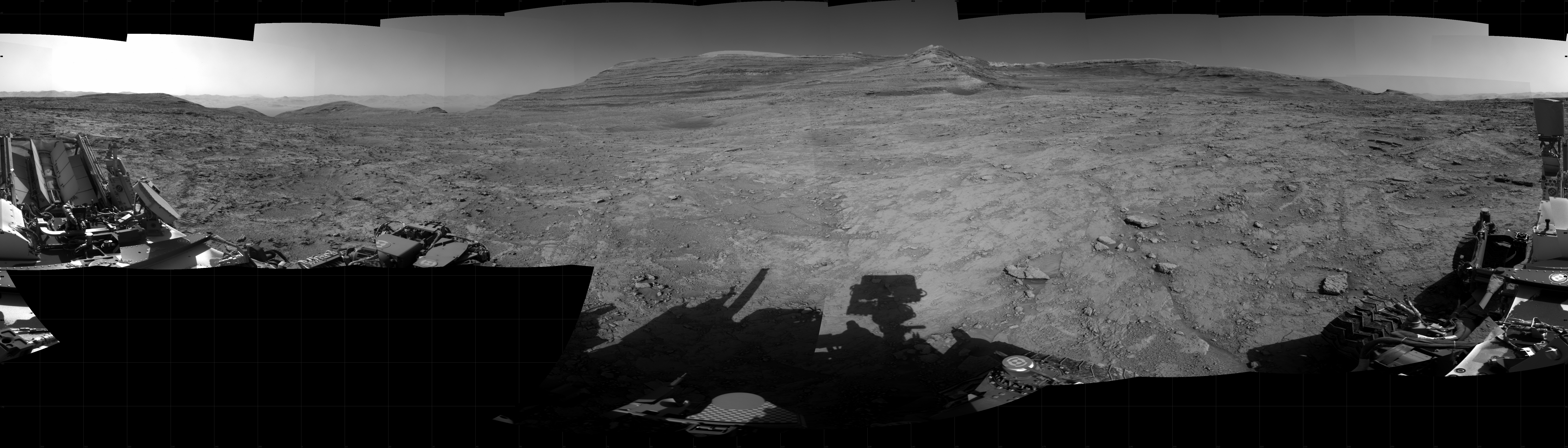 Panoramic black-and-white image of the rocky Martian landscape taken by the Curiosity rover, showing rugged terrain, layered hills in the distance, and parts of the rover's structure in the foreground.