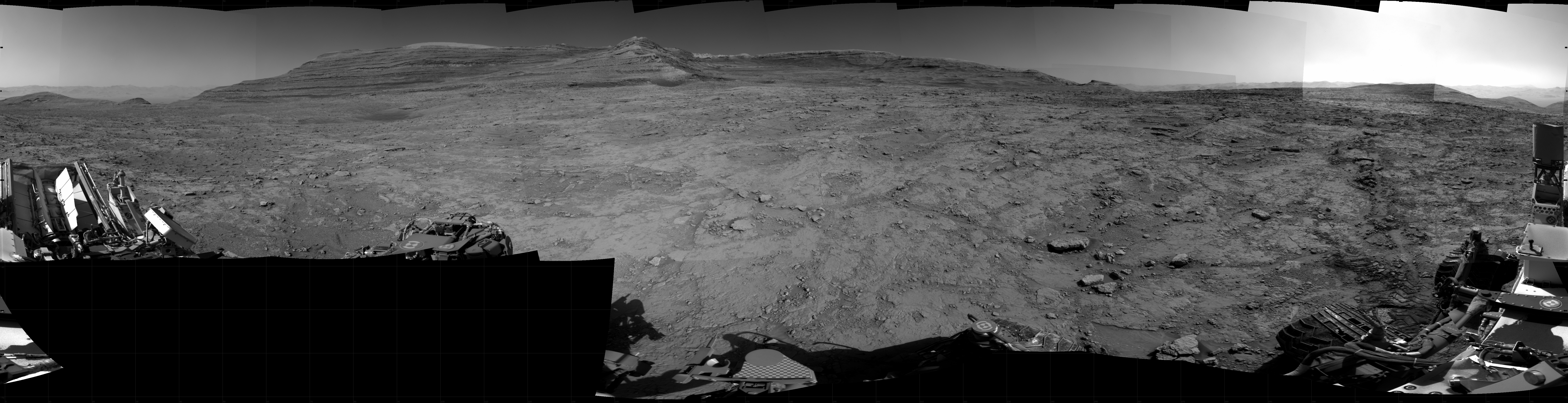 Panoramic black-and-white image of the rocky Martian landscape taken by the Curiosity rover, showing rugged terrain, layered hills in the distance, and parts of the rover's structure in the foreground.