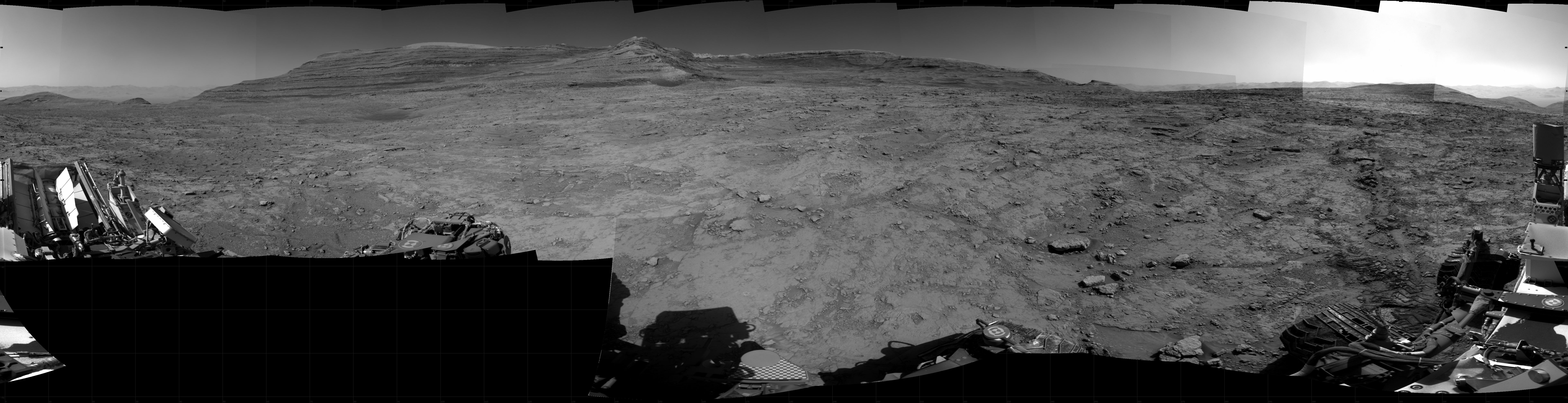 Panoramic black-and-white image of the rocky Martian landscape taken by the Curiosity rover, showing rugged terrain, layered hills in the distance, and parts of the rover's structure in the foreground.