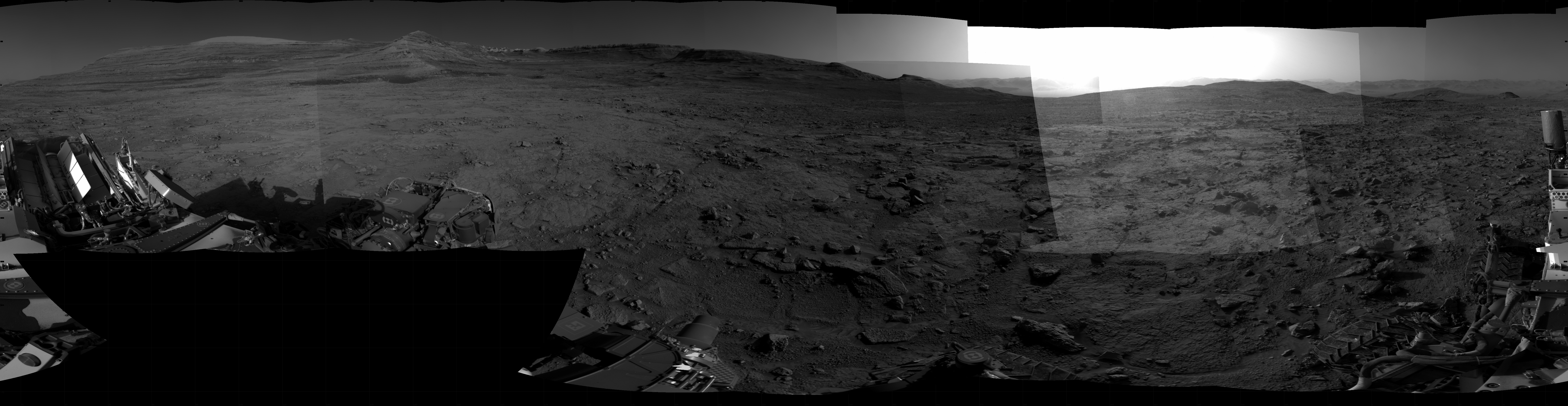 Panoramic black-and-white image of the rocky Martian landscape taken by the Curiosity rover, showing rugged terrain, layered hills in the distance, and parts of the rover's structure in the foreground.