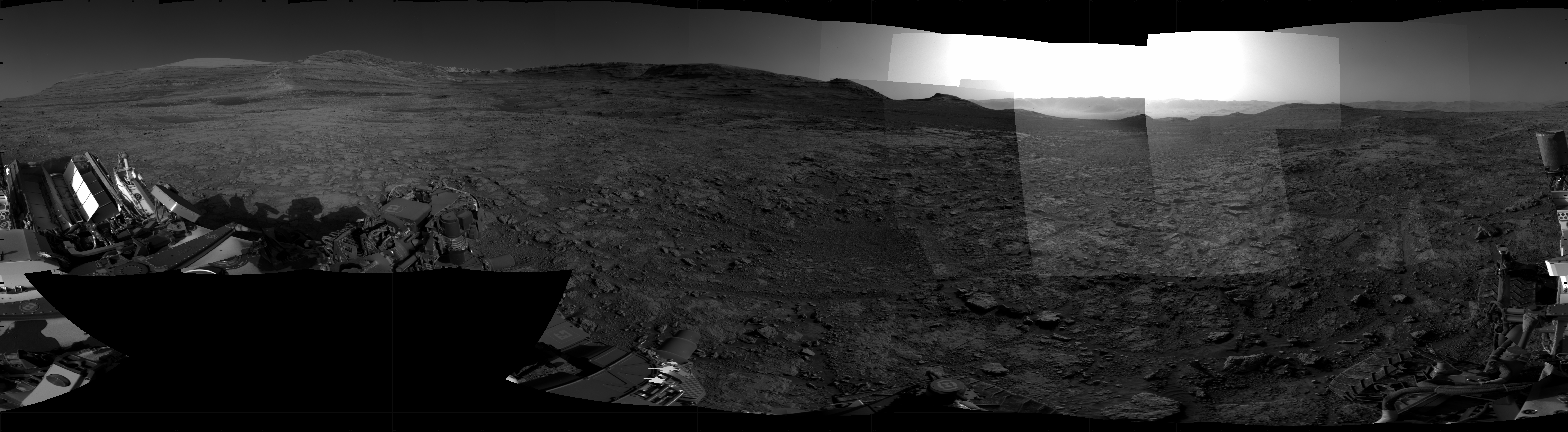Panoramic black-and-white image of the rocky Martian landscape taken by the Curiosity rover, showing rugged terrain, layered hills in the distance, and parts of the rover's structure in the foreground.