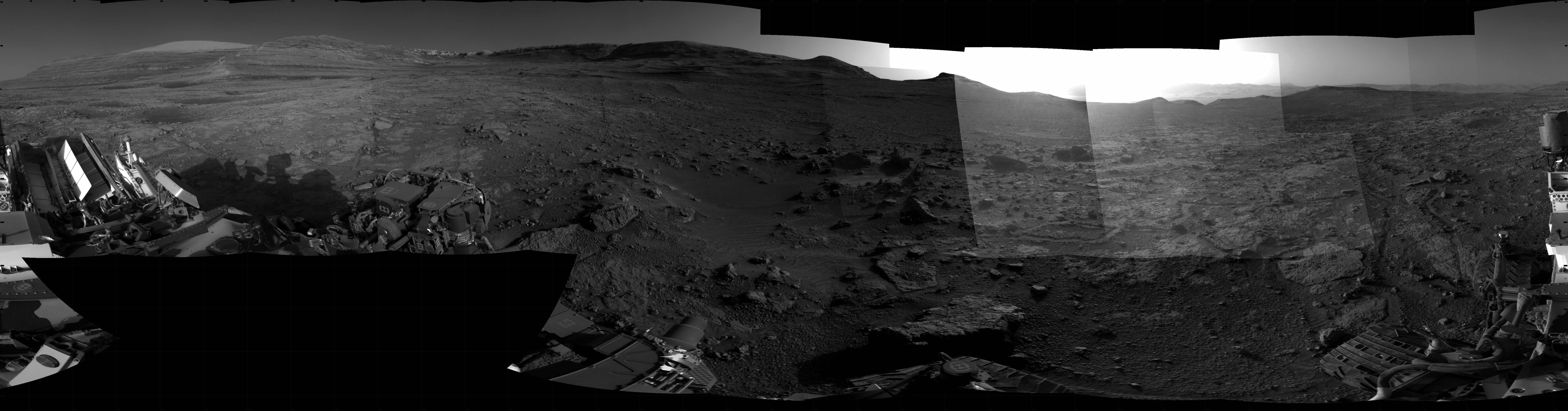 Panoramic black-and-white image of the rocky Martian landscape taken by the Curiosity rover, showing rugged terrain, layered hills in the distance, and parts of the rover's structure in the foreground.