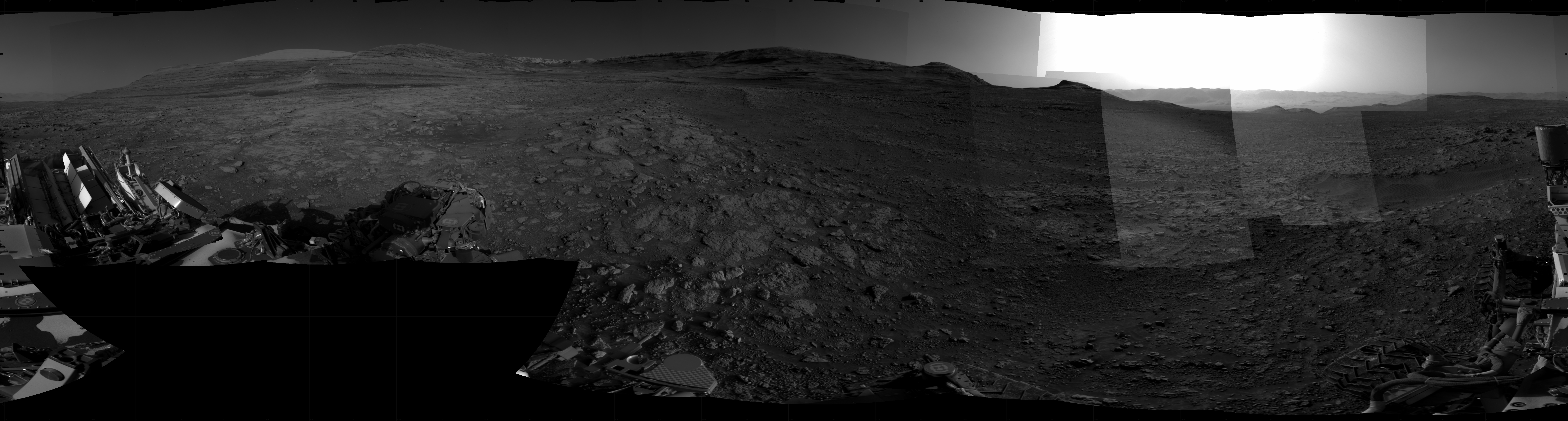 Panoramic black-and-white image of the rocky Martian landscape taken by the Curiosity rover, showing rugged terrain, layered hills in the distance, and parts of the rover's structure in the foreground.