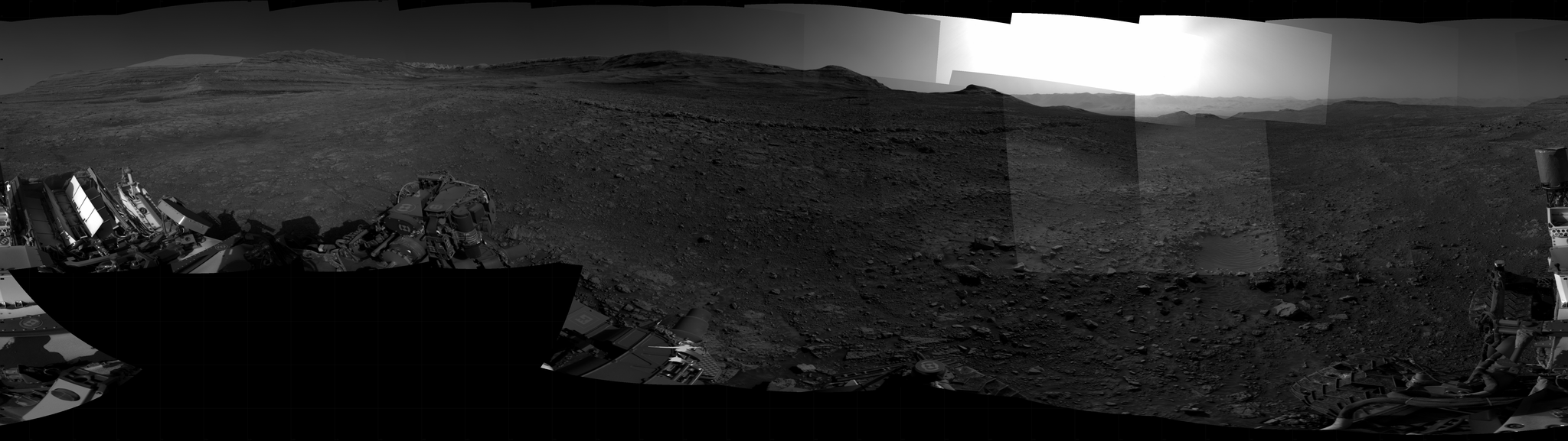Panoramic black-and-white image of the rocky Martian landscape taken by the Curiosity rover, showing rugged terrain, layered hills in the distance, and parts of the rover's structure in the foreground.