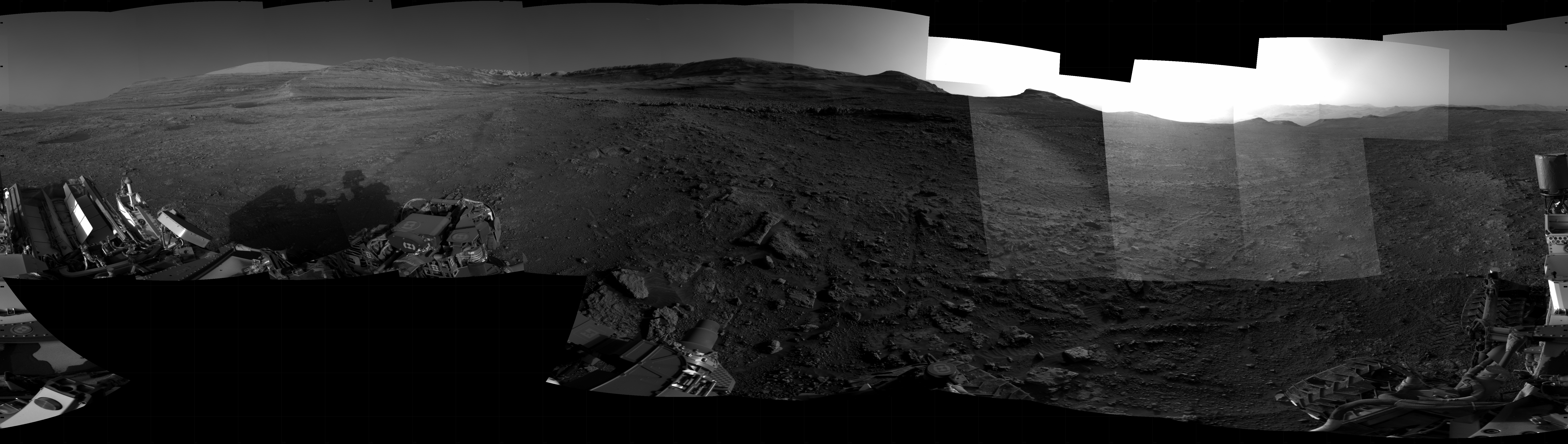 Panoramic black-and-white image of the rocky Martian landscape taken by the Curiosity rover, showing rugged terrain, layered hills in the distance, and parts of the rover's structure in the foreground.
