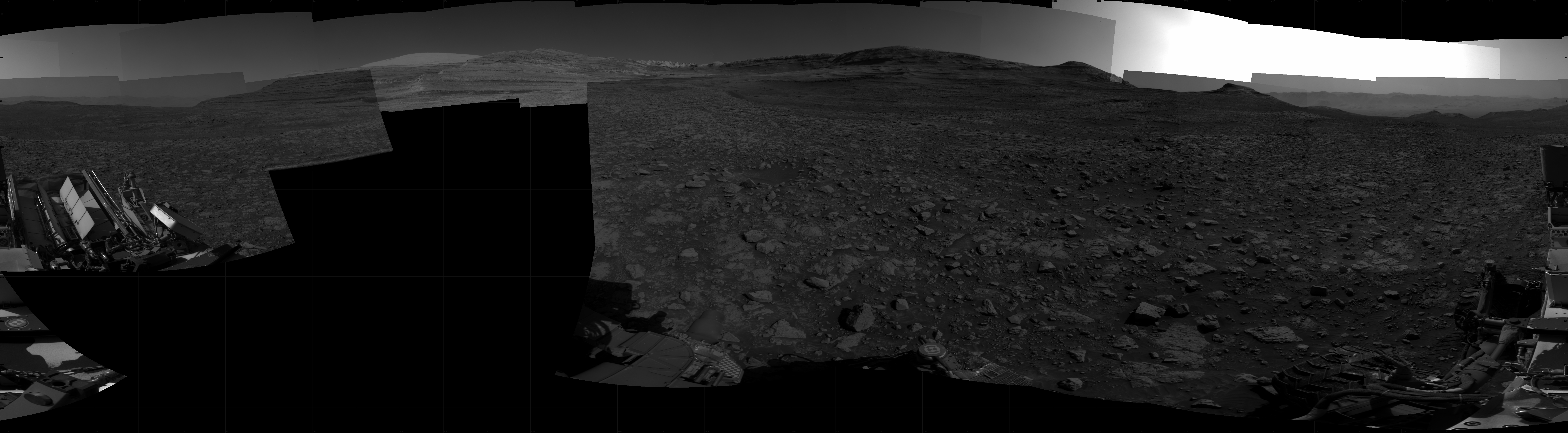 Panoramic black-and-white image of the rocky Martian landscape taken by the Curiosity rover, showing rugged terrain, layered hills in the distance, and parts of the rover's structure in the foreground.