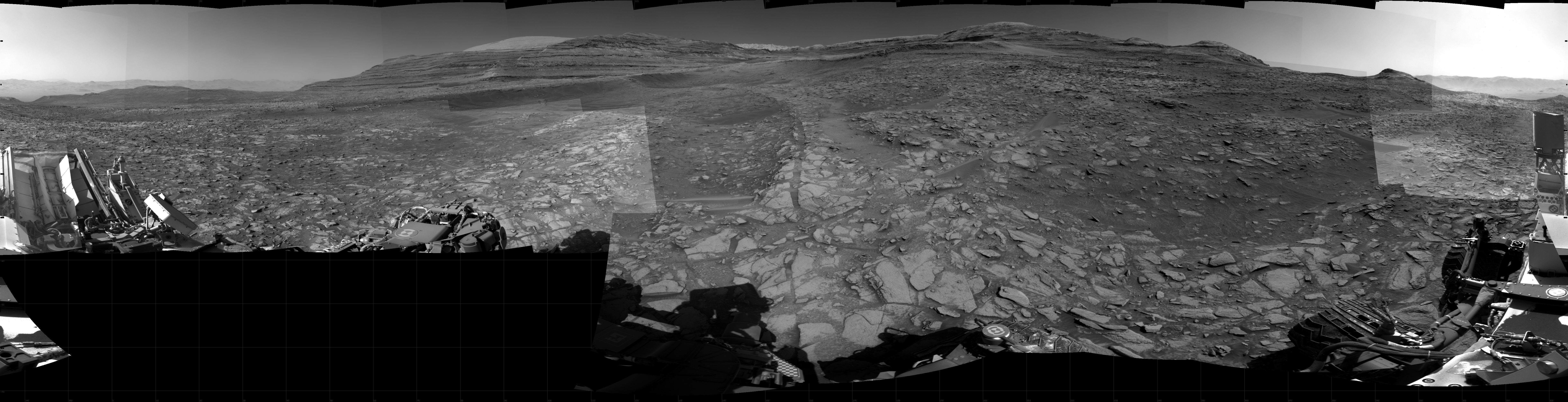 Panoramic black-and-white image of the rocky Martian landscape taken by the Curiosity rover, showing rugged terrain, layered hills in the distance, and parts of the rover's structure in the foreground.