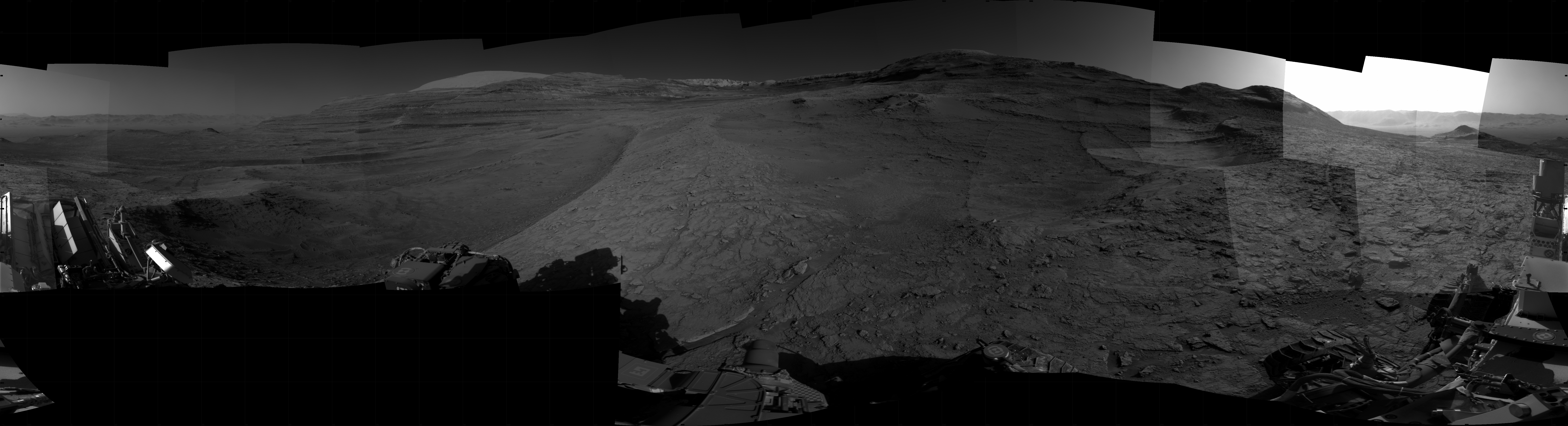 Panoramic black-and-white image of the rocky Martian landscape taken by the Curiosity rover, showing rugged terrain, layered hills in the distance, and parts of the rover's structure in the foreground.
