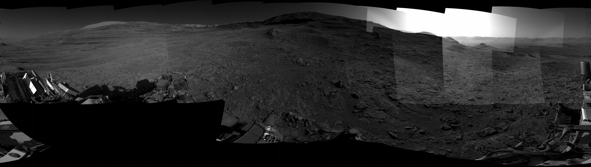 Panoramic black-and-white image of the rocky Martian landscape taken by the Curiosity rover, showing rugged terrain, layered hills in the distance, and parts of the rover's structure in the foreground.