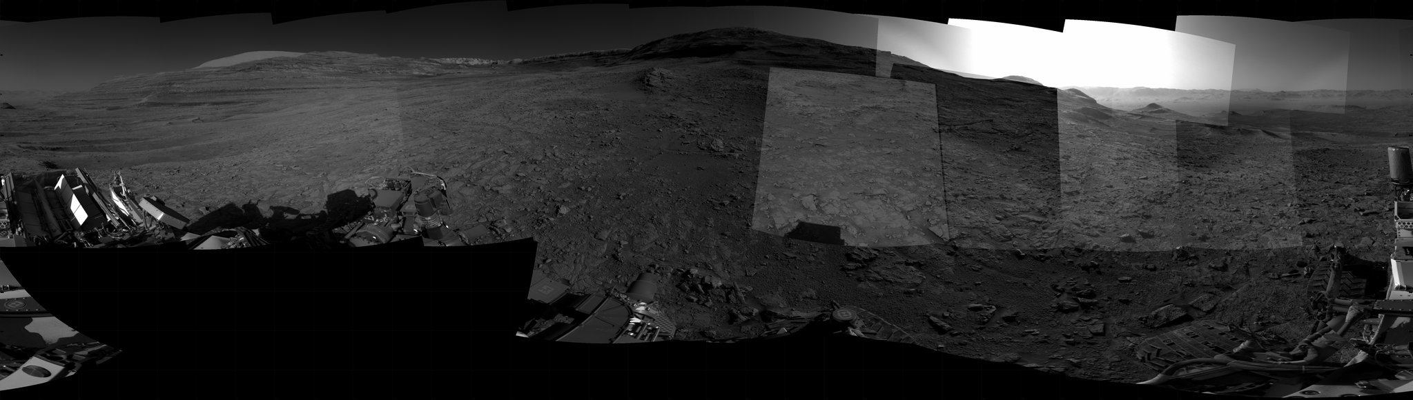 Panoramic black-and-white image of the rocky Martian landscape taken by the Curiosity rover, showing rugged terrain, layered hills in the distance, and parts of the rover's structure in the foreground.Panoramic black-and-white image of the rocky Martian landscape taken by the Curiosity rover, showing rugged terrain, layered hills in the distance, and parts of the rover's structure in the foreground.