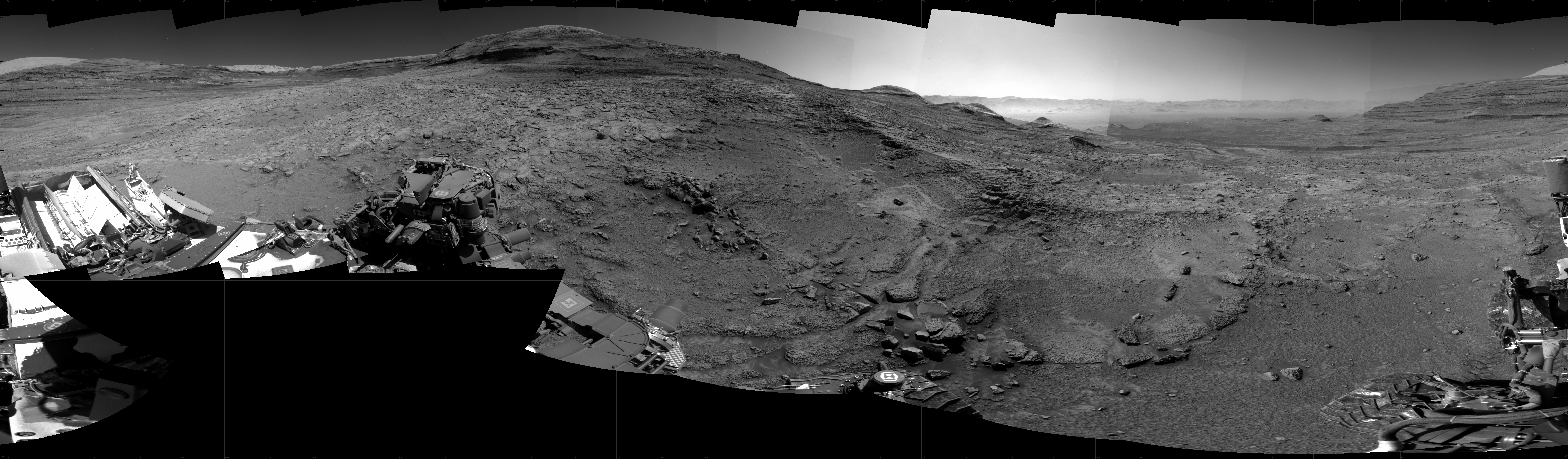 Panoramic black-and-white image of the rocky Martian landscape taken by the Curiosity rover, showing rugged terrain, layered hills in the distance, and parts of the rover's structure in the foreground.