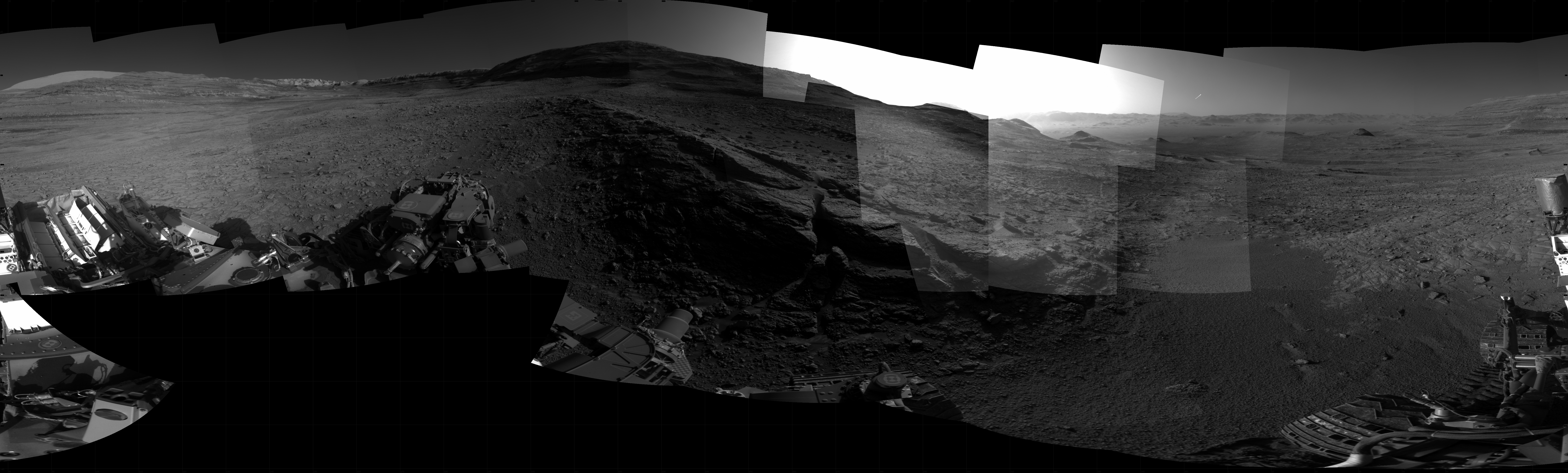 Panoramic black-and-white image of the rocky Martian landscape taken by the Curiosity rover, showing rugged terrain, layered hills in the distance, and parts of the rover's structure in the foreground.