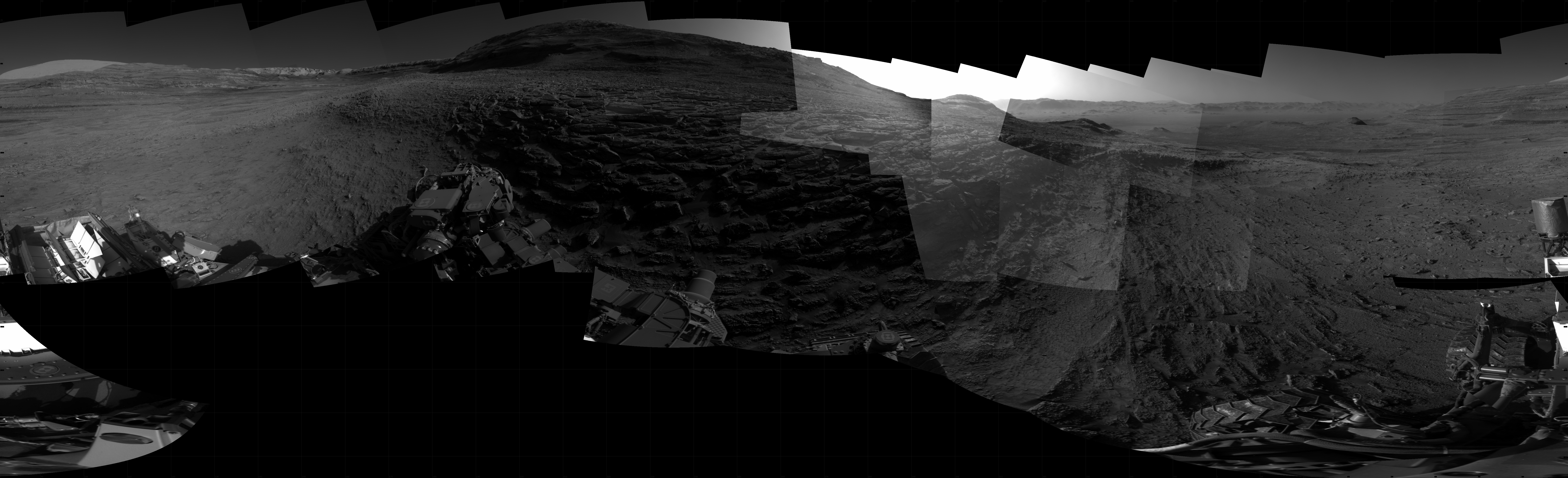 Panoramic black-and-white image of the rocky Martian landscape taken by the Curiosity rover, showing rugged terrain, layered hills in the distance, and parts of the rover's structure in the foreground.