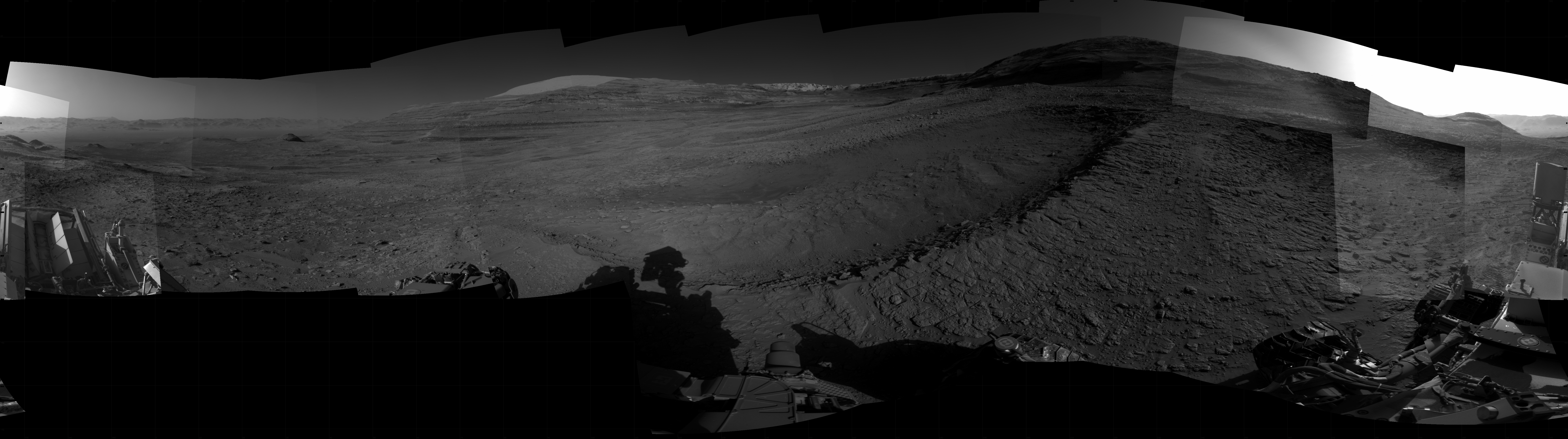 Panoramic black-and-white image of the rocky Martian landscape taken by the Curiosity rover, showing rugged terrain, layered hills in the distance, and parts of the rover's structure in the foreground.
