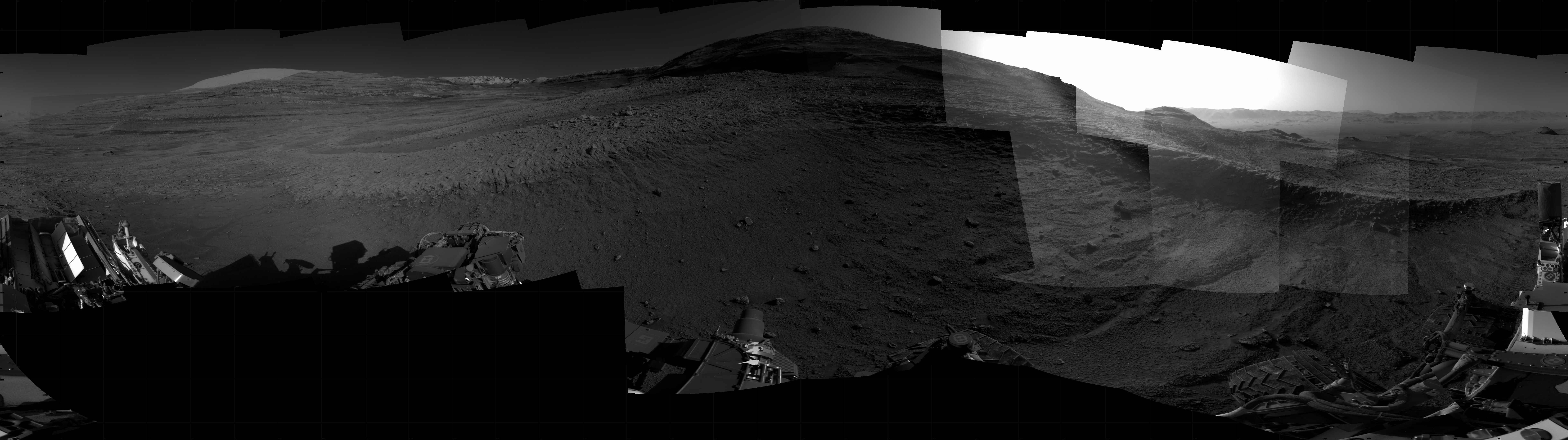Panoramic black-and-white image of the rocky Martian landscape taken by the Curiosity rover, showing rugged terrain, layered hills in the distance, and parts of the rover's structure in the foreground.