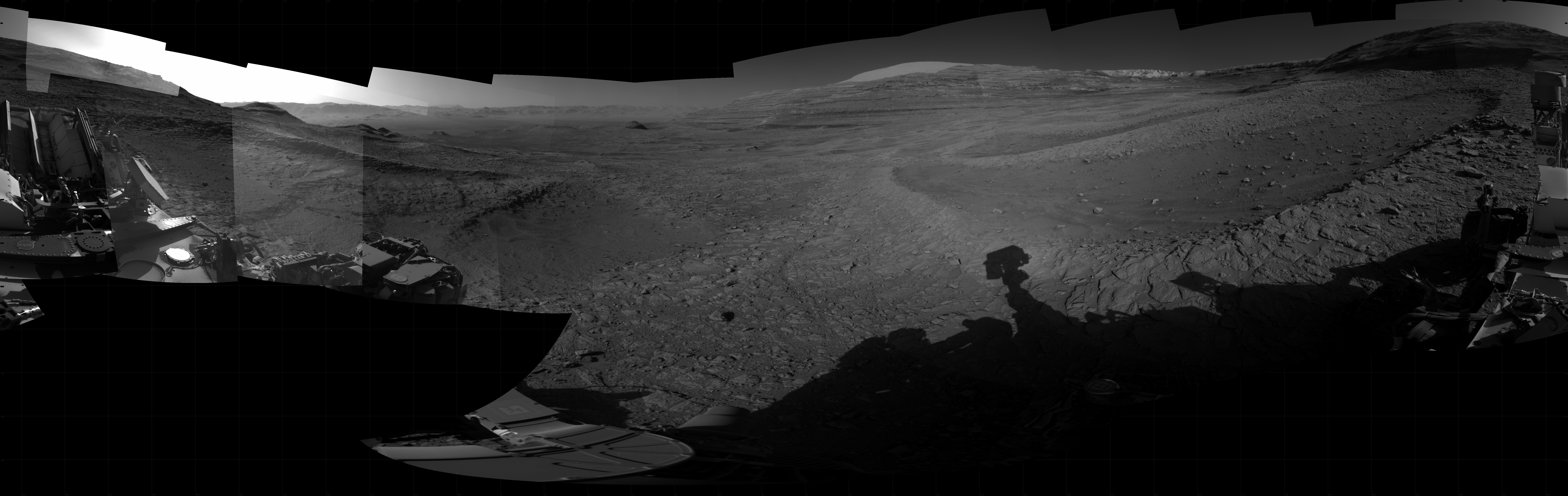 Panoramic black-and-white image of the rocky Martian landscape taken by the Curiosity rover, showing rugged terrain, layered hills in the distance, and parts of the rover's structure in the foreground.