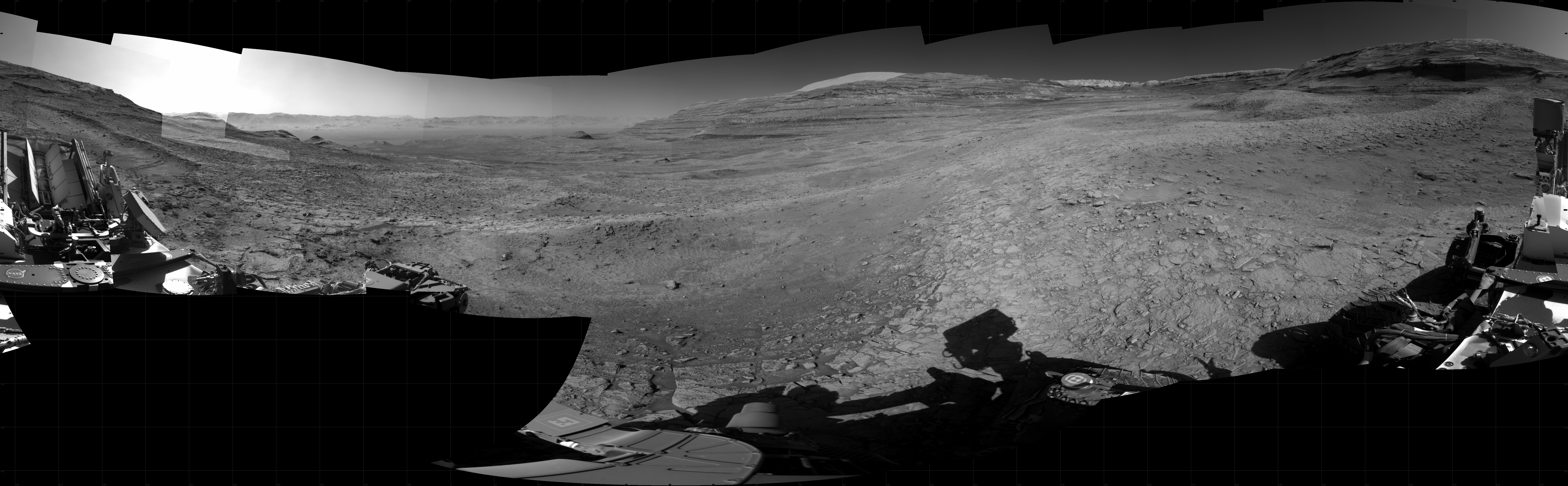 Panoramic black-and-white image of the rocky Martian landscape taken by the Curiosity rover, showing rugged terrain, layered hills in the distance, and parts of the rover's structure in the foreground.