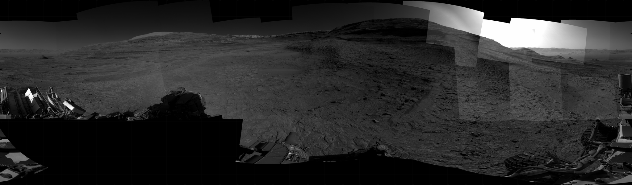 Panoramic black-and-white image of the rocky Martian landscape taken by the Curiosity rover, showing rugged terrain, layered hills in the distance, and parts of the rover's structure in the foreground.