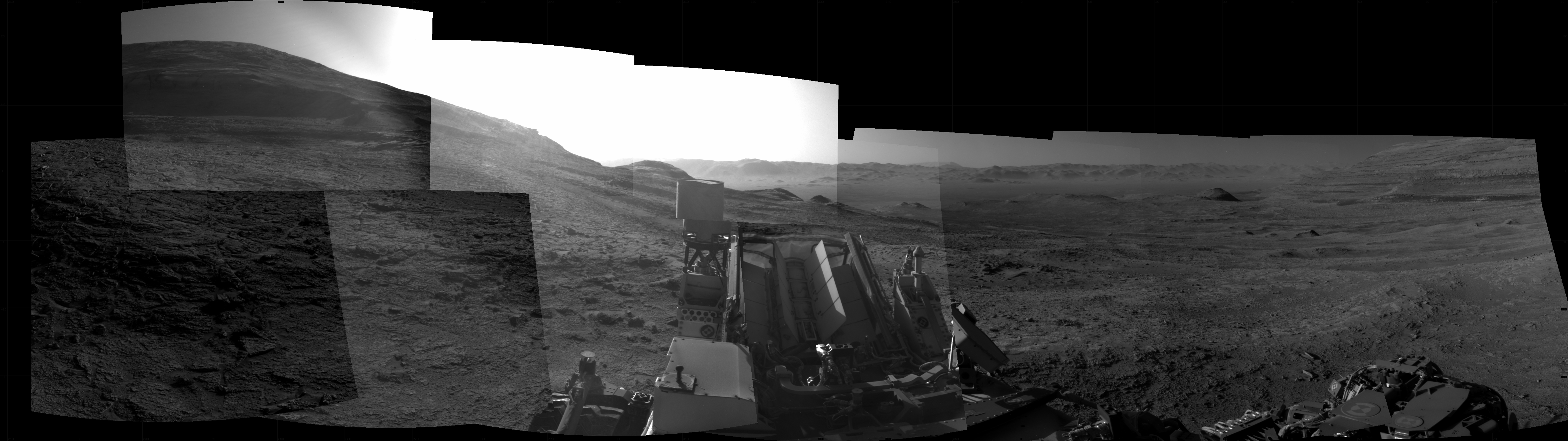Panoramic black-and-white image of the rocky Martian landscape taken by the Curiosity rover, showing rugged terrain, layered hills in the distance, and parts of the rover's structure in the foreground.