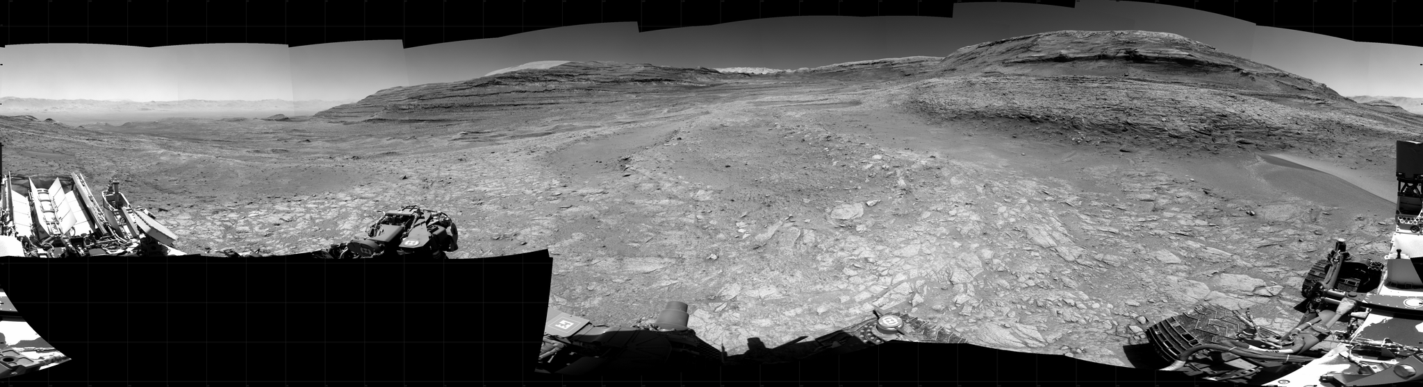Panoramic black-and-white image of the rocky Martian landscape taken by the Curiosity rover, showing rugged terrain, layered hills in the distance, and parts of the rover's structure in the foreground.