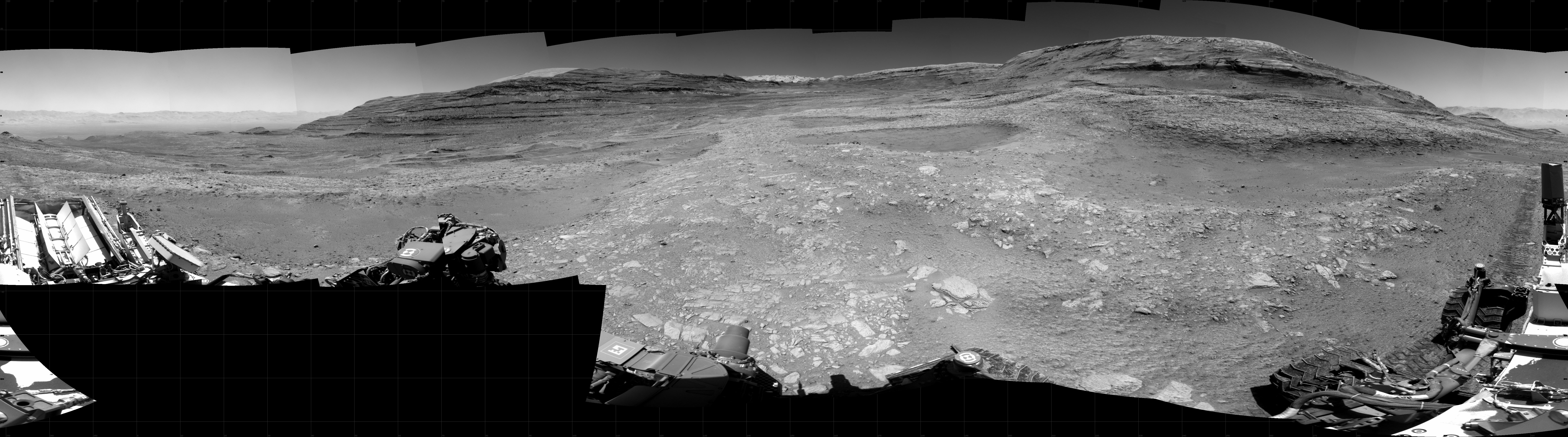 Panoramic black-and-white image of the rocky Martian landscape taken by the Curiosity rover, showing rugged terrain, layered hills in the distance, and parts of the rover's structure in the foreground.