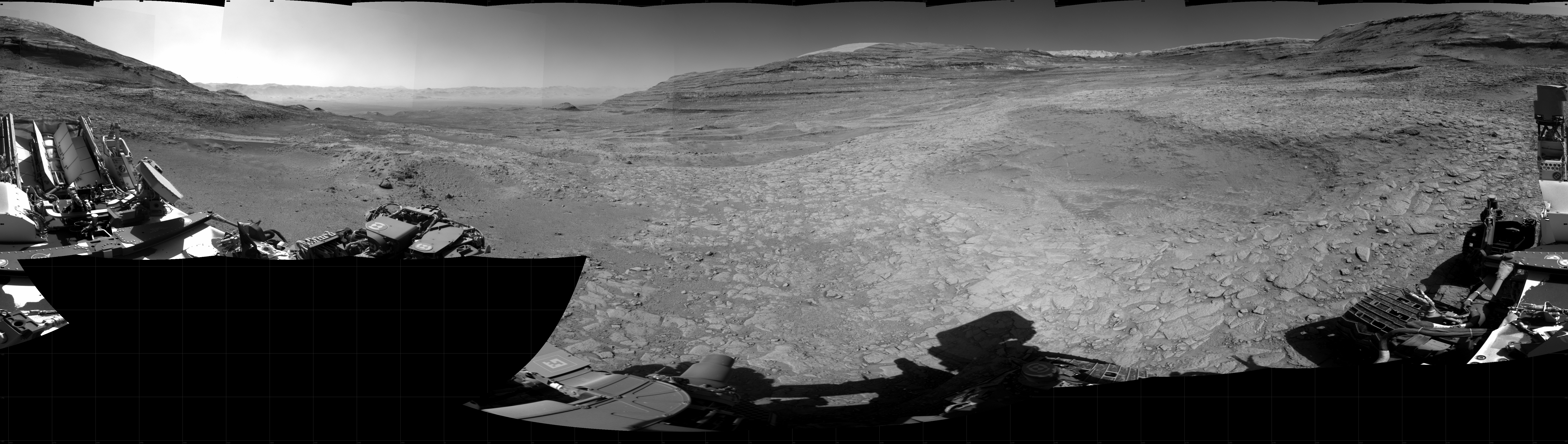 Panoramic black-and-white image of the rocky Martian landscape taken by the Curiosity rover, showing rugged terrain, layered hills in the distance, and parts of the rover's structure in the foreground.