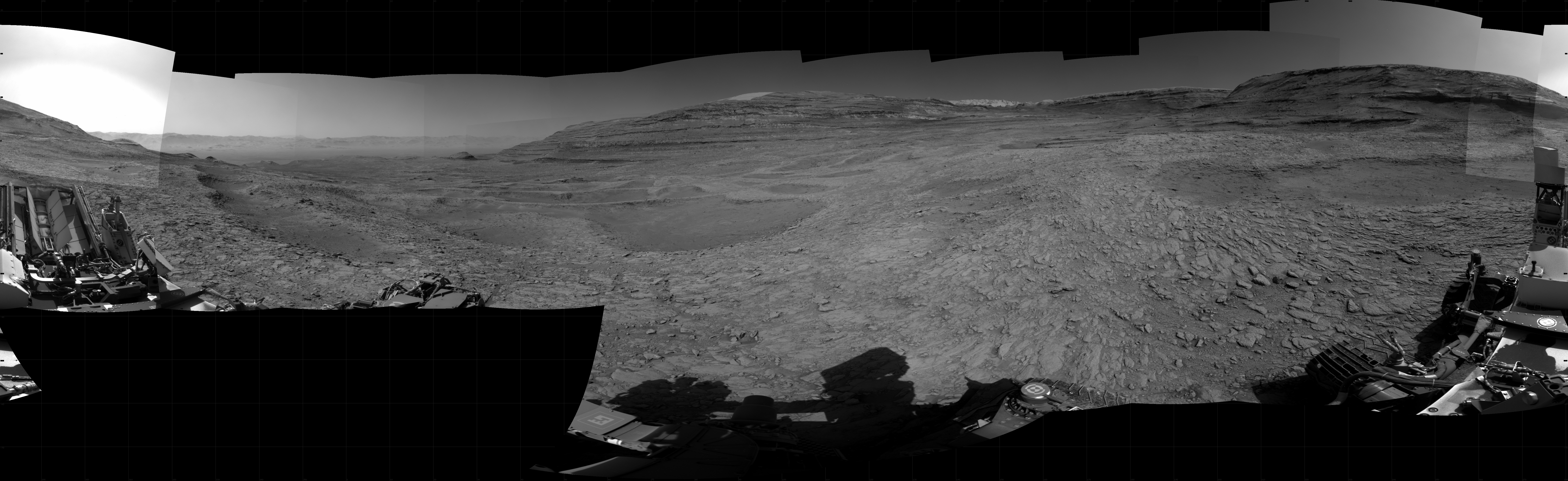 Panoramic black-and-white image of the rocky Martian landscape taken by the Curiosity rover, showing rugged terrain, layered hills in the distance, and parts of the rover's structure in the foreground.