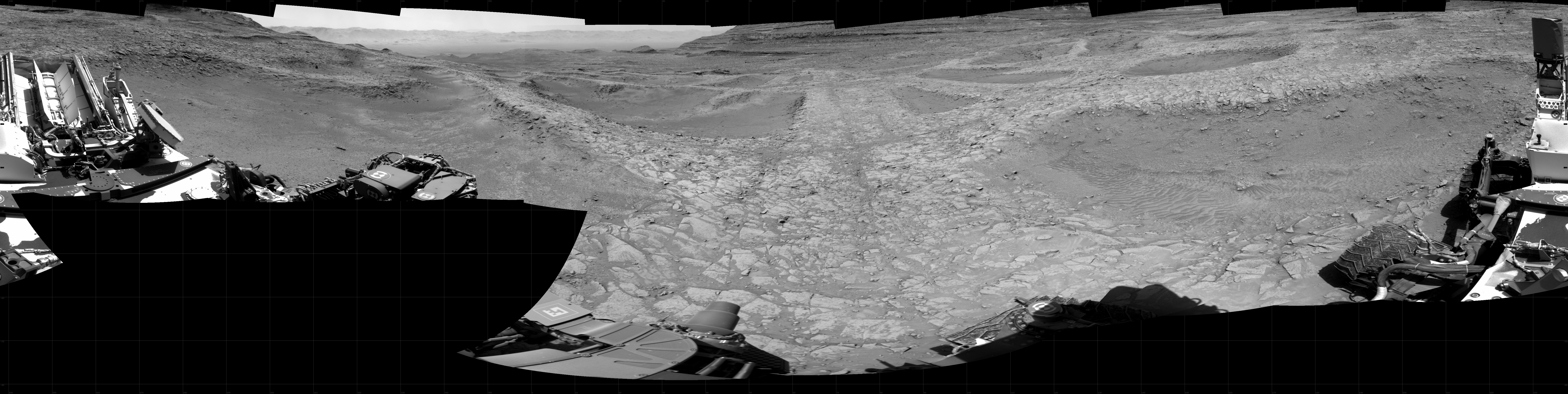 Panoramic black-and-white image of the rocky Martian landscape taken by the Curiosity rover, showing rugged terrain, layered hills in the distance, and parts of the rover's structure in the foreground.