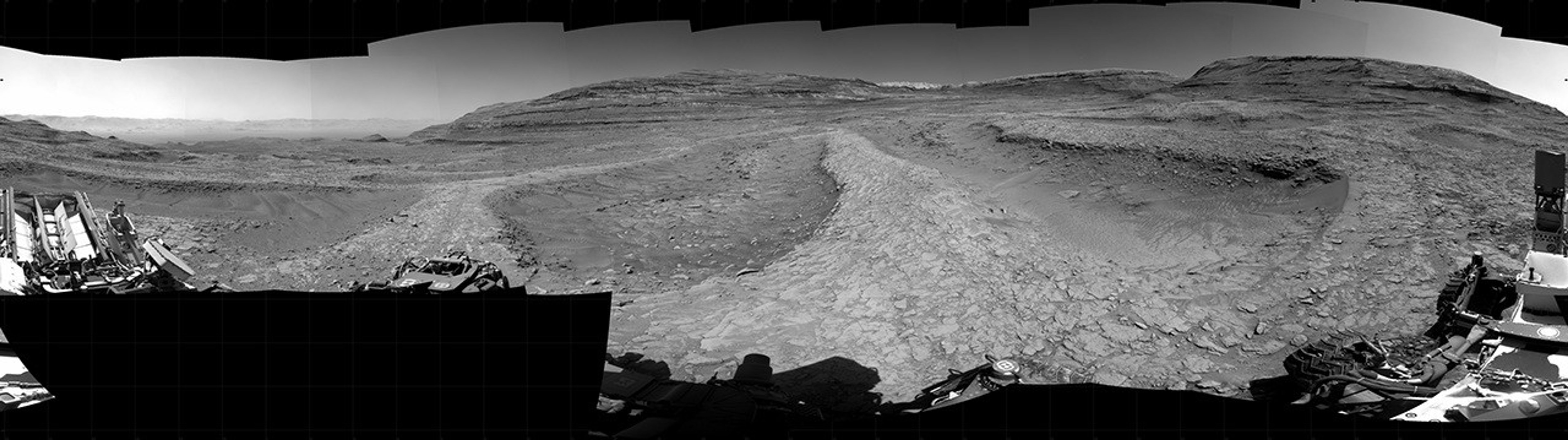 Panoramic black-and-white image of the rocky Martian landscape taken by the Curiosity rover, showing rugged terrain, layered hills in the distance, and parts of the rover's structure in the foreground.