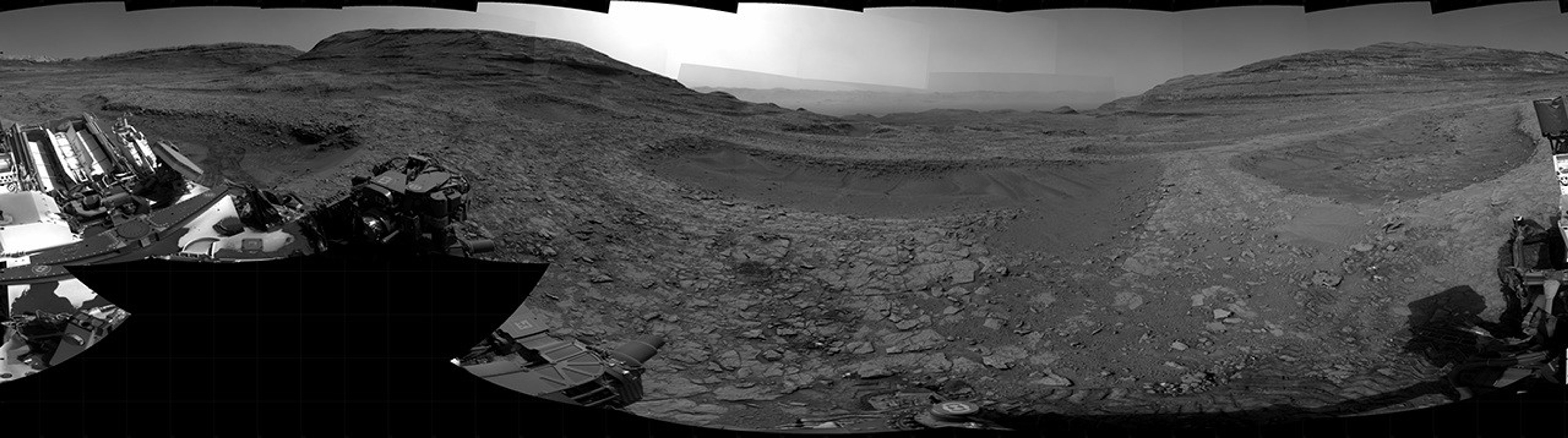 Panoramic black-and-white image of the rocky Martian landscape taken by the Curiosity rover, showing rugged terrain, layered hills in the distance, and parts of the rover's structure in the foreground.
