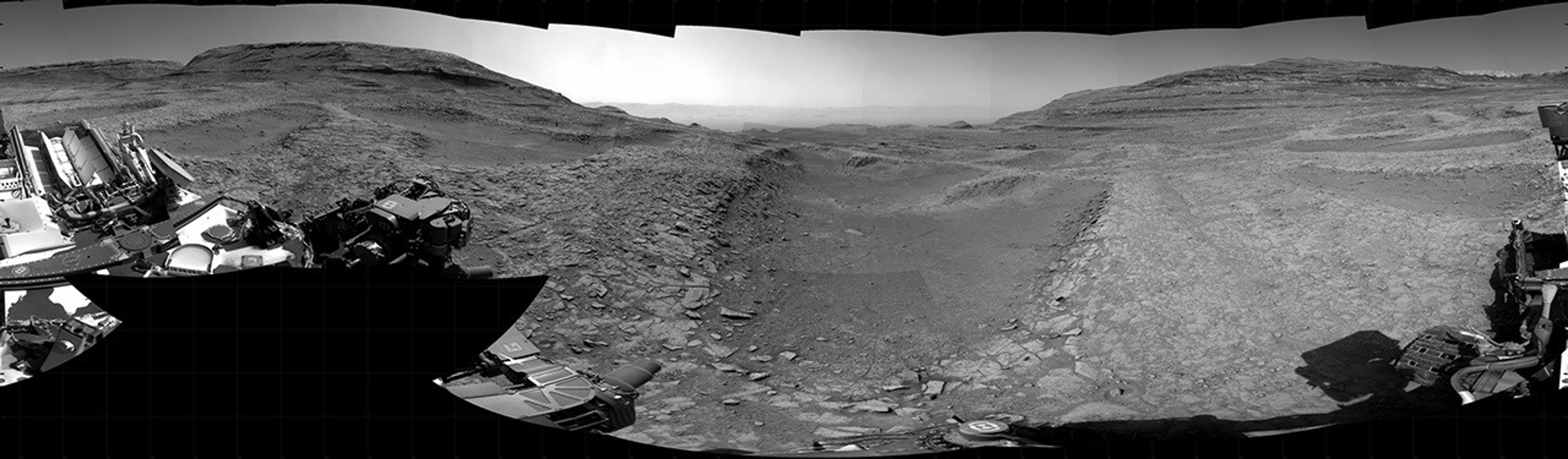 Panoramic black-and-white image of the rocky Martian landscape taken by the Curiosity rover, showing rugged terrain, layered hills in the distance, and parts of the rover's structure in the foreground.