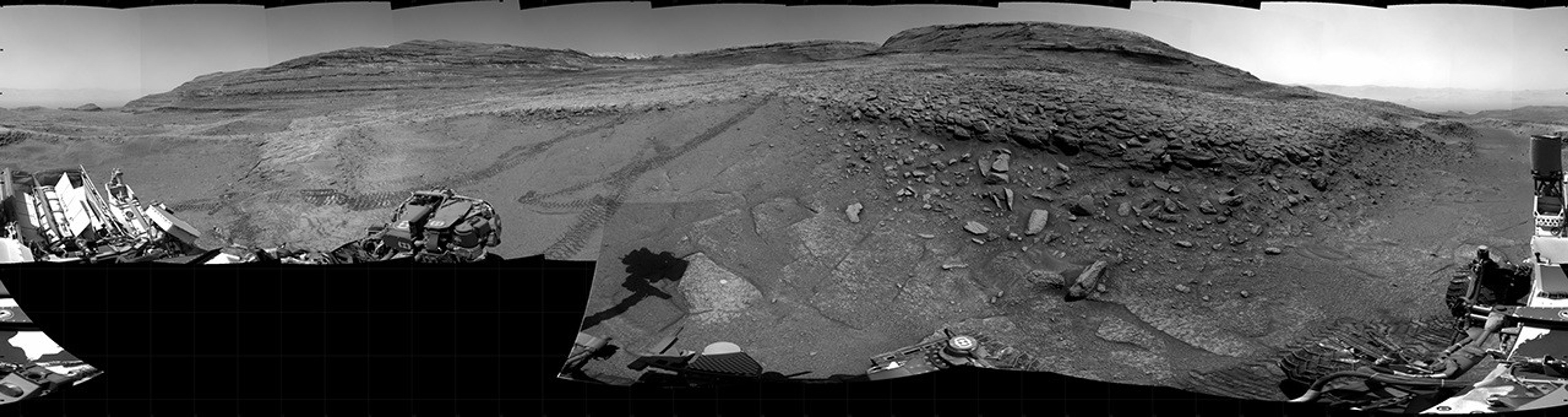 Panoramic black-and-white image of the rocky Martian landscape taken by the Curiosity rover, showing rugged terrain, layered hills in the distance, and parts of the rover's structure in the foreground.