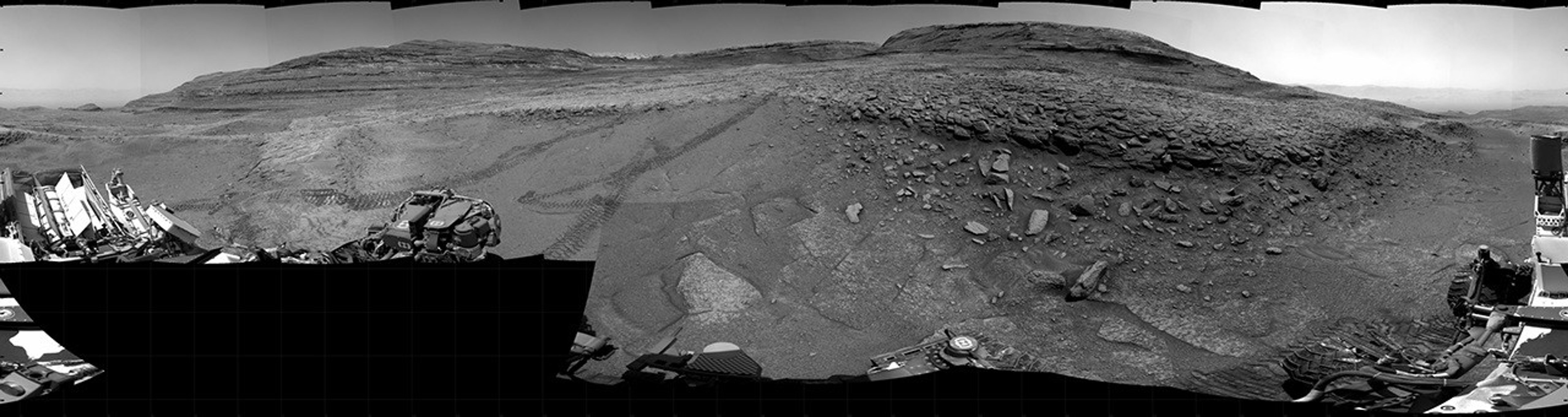 Panoramic black-and-white image of the rocky Martian landscape taken by the Curiosity rover, showing rugged terrain, layered hills in the distance, and parts of the rover's structure in the foreground.
