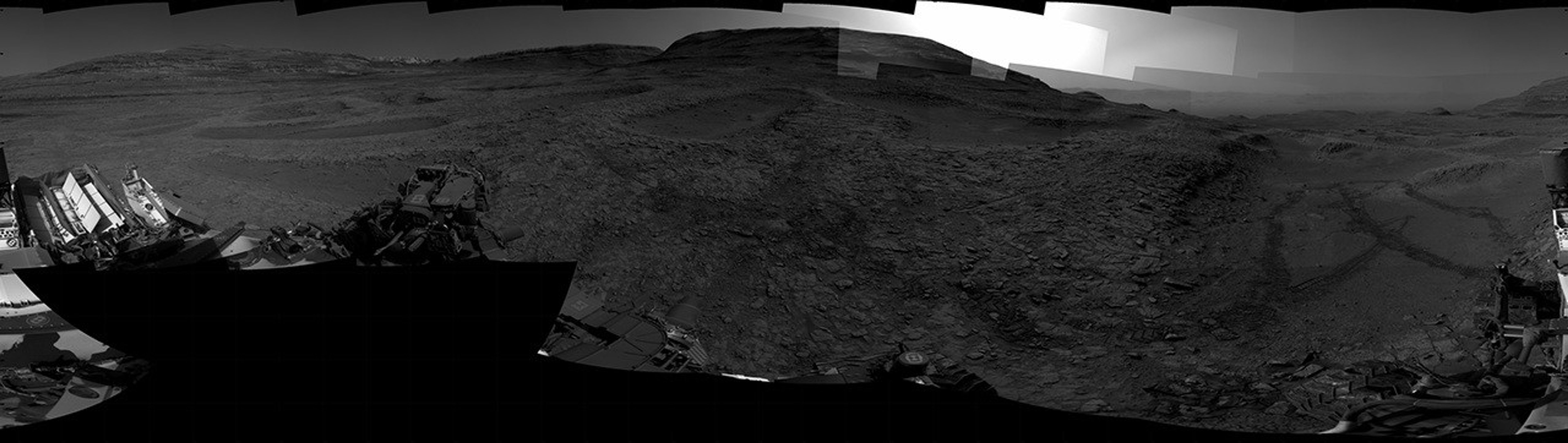 A grayscale panorama of the Martian surface, possibly at night or in twilight, shows a wide field of flat, dark gray terrain dotted with flat, angular, medium-sized rocks stretching into the distance where features rise from the ground. At the far right of the frame, a very bright light source illuminates the sky above several small hills. Portions of the Curiosity rover are visible along the bottom of the image.