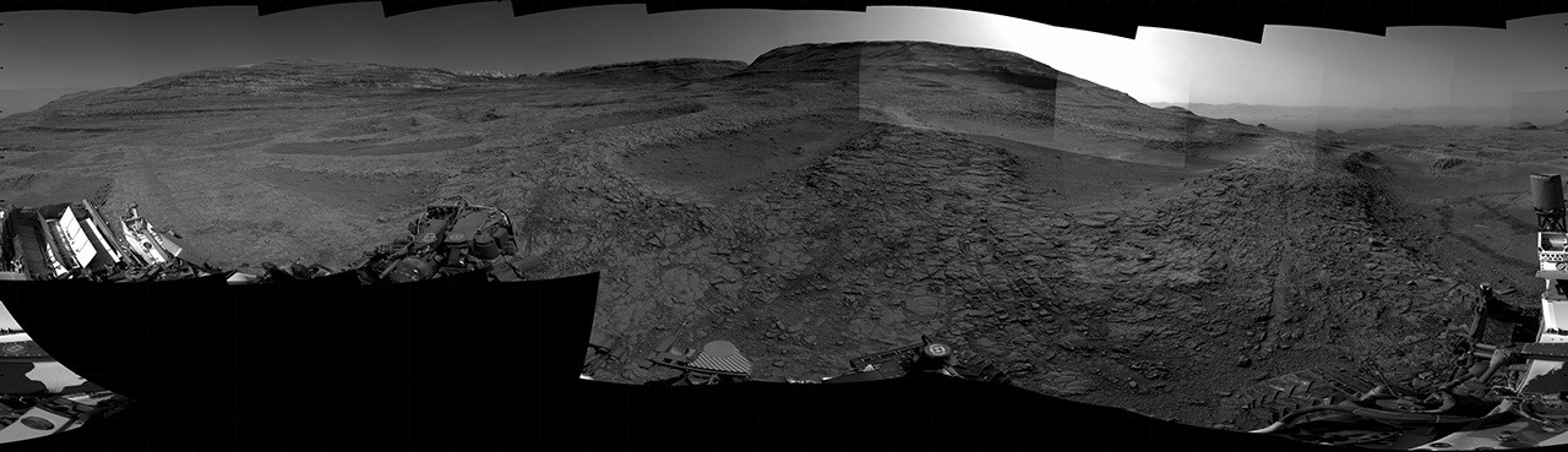 A grayscale panorama of the Martian surface, possibly at night or in twilight, shows a wide field of flat, dark gray terrain dotted with multiple jagged, medium-sized rocks stretching into the distance, where hills rise from the ground on the horizon. At center-right, a bright light behind a hill illuminates part of the sky, while the rest of the scene is fairly dark. Portions of the Curiosity rover are visible along the bottom of the image, including wheels in the lower-right corner.