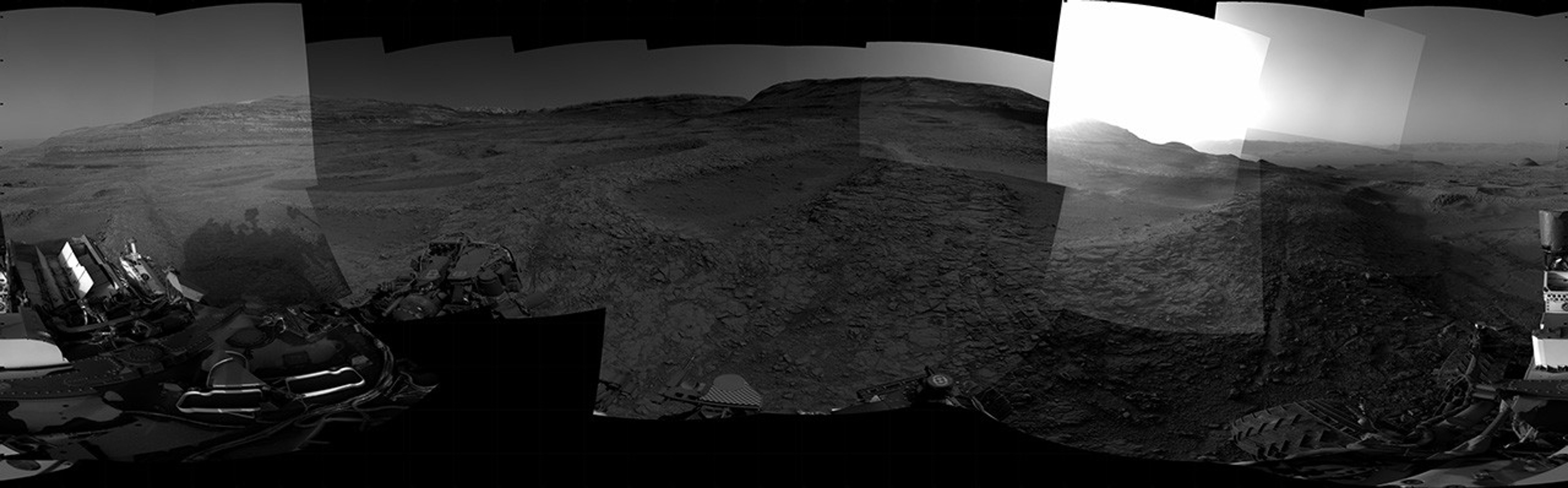 A grayscale panorama of the Martian surface, possibly at night or in twilight, shows a wide field of flat, dark gray terrain dotted with multiple jagged, medium-sized rocks stretching into the distance, where hills rise from the ground on the horizon. At center-right, a bright light behind a hill illuminates part of the sky, while the rest of the scene is fairly dark. Portions of the Curiosity rover are visible along the bottom of the image, including wheels in the lower-right corner.